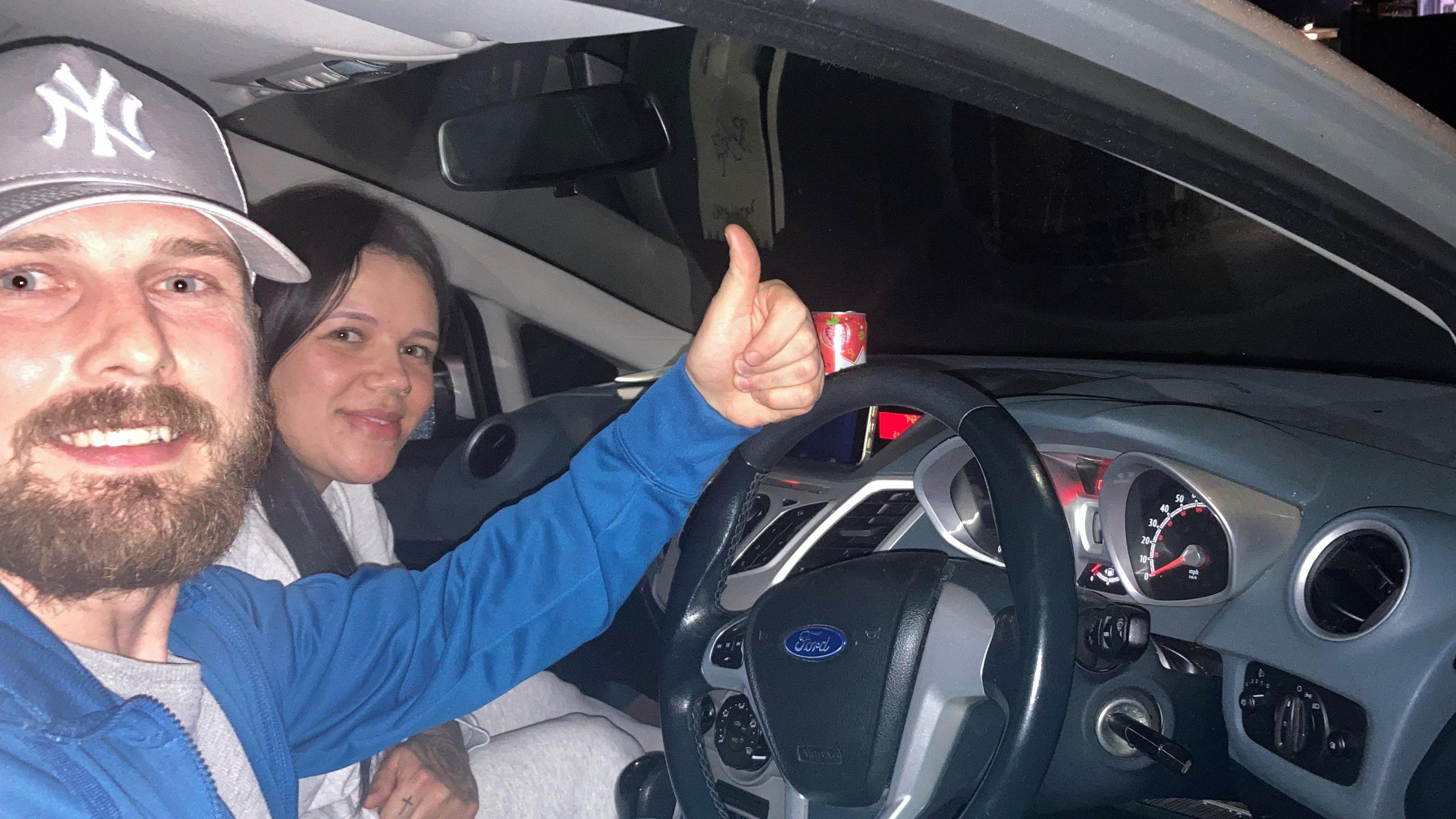 Two people smiling and sitting in their car with a thumbs up. 