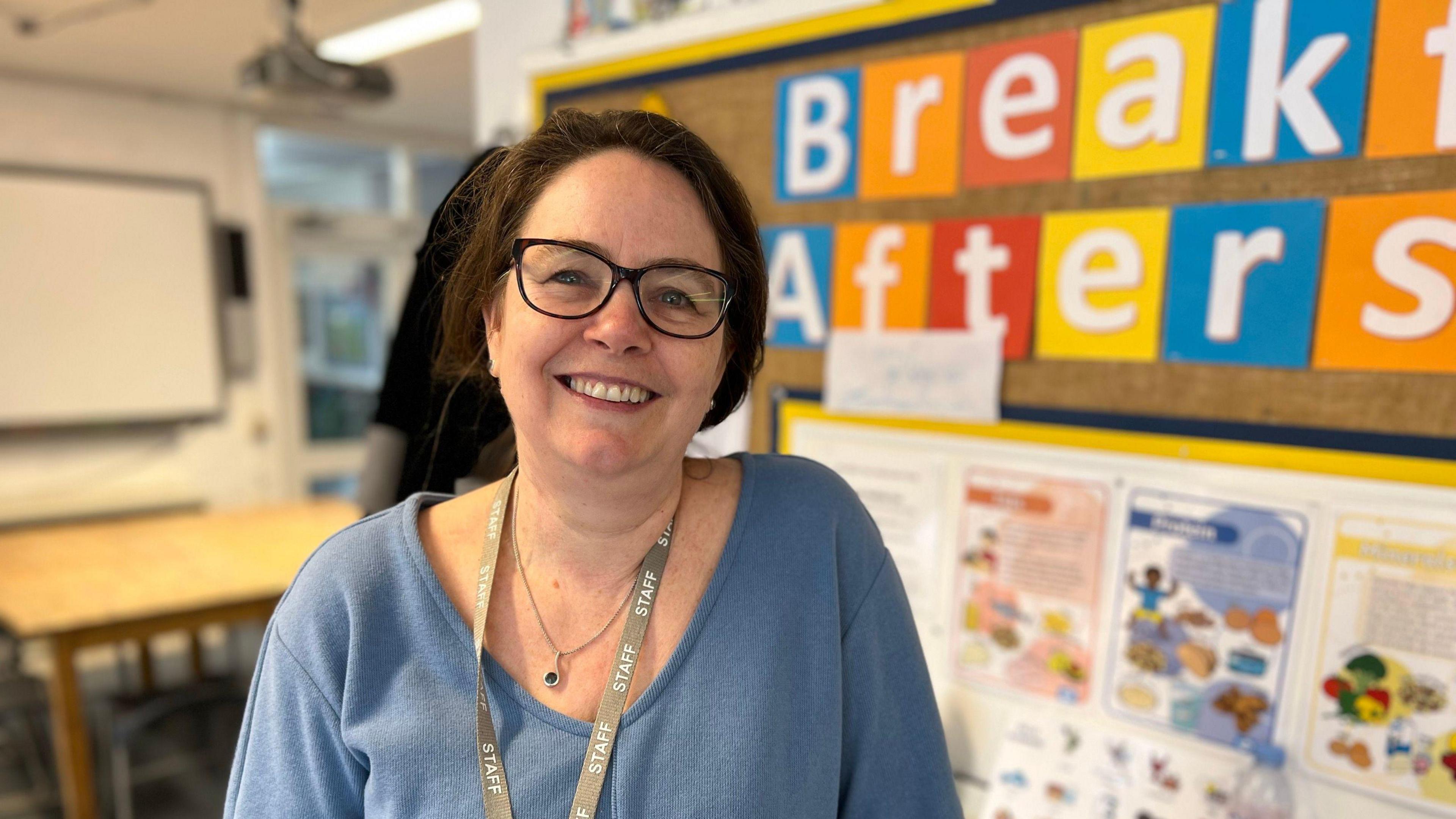 Jess Tweedie stands in front of a noticeboard in a classroom. She has short brown hair and wears glasses. She wears a light blue top and has a staff lanyard around her neck.