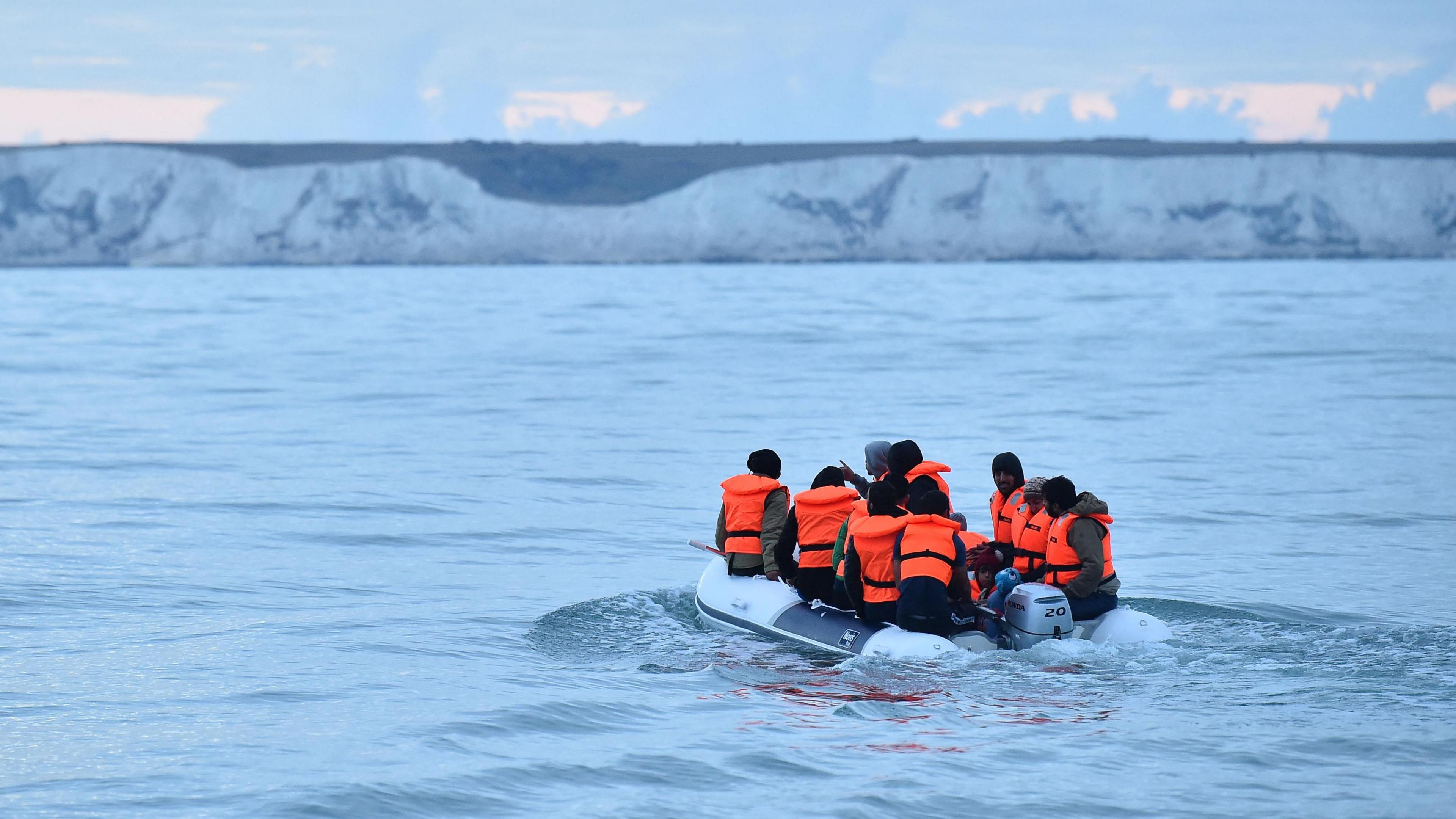 A group of people in orange life jackets on a small boat in a large body of water travelling towards white cliffs which are in the background.