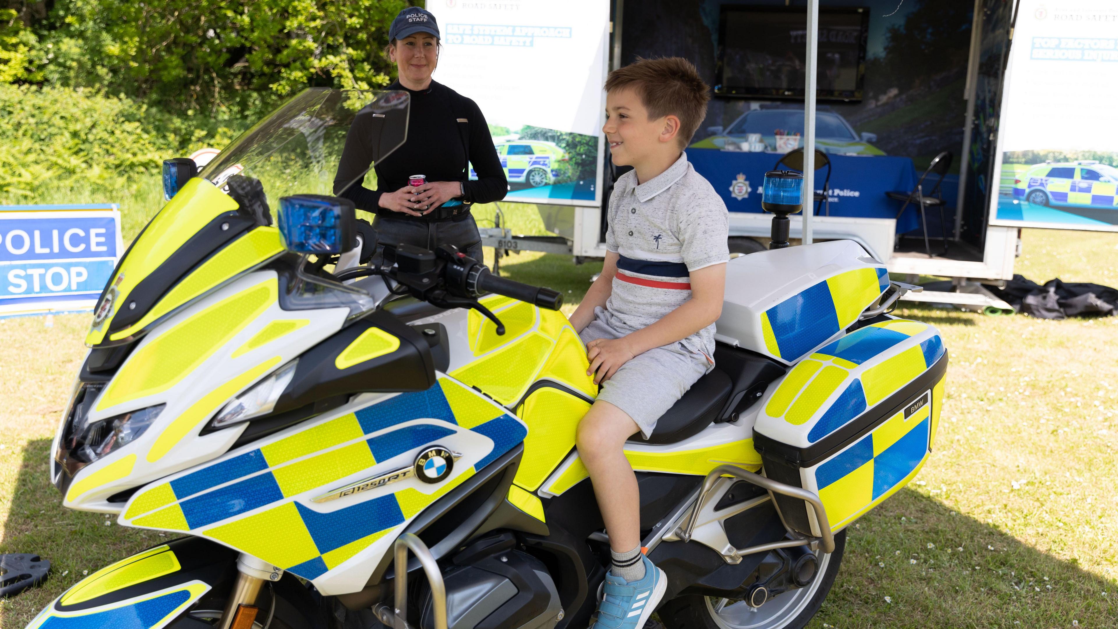 A young boy wearing grey shorts and a t-shirt smiling and sitting on a police motorcycle while a policewoman wearing black stands about a foot away, smiling at the boy.