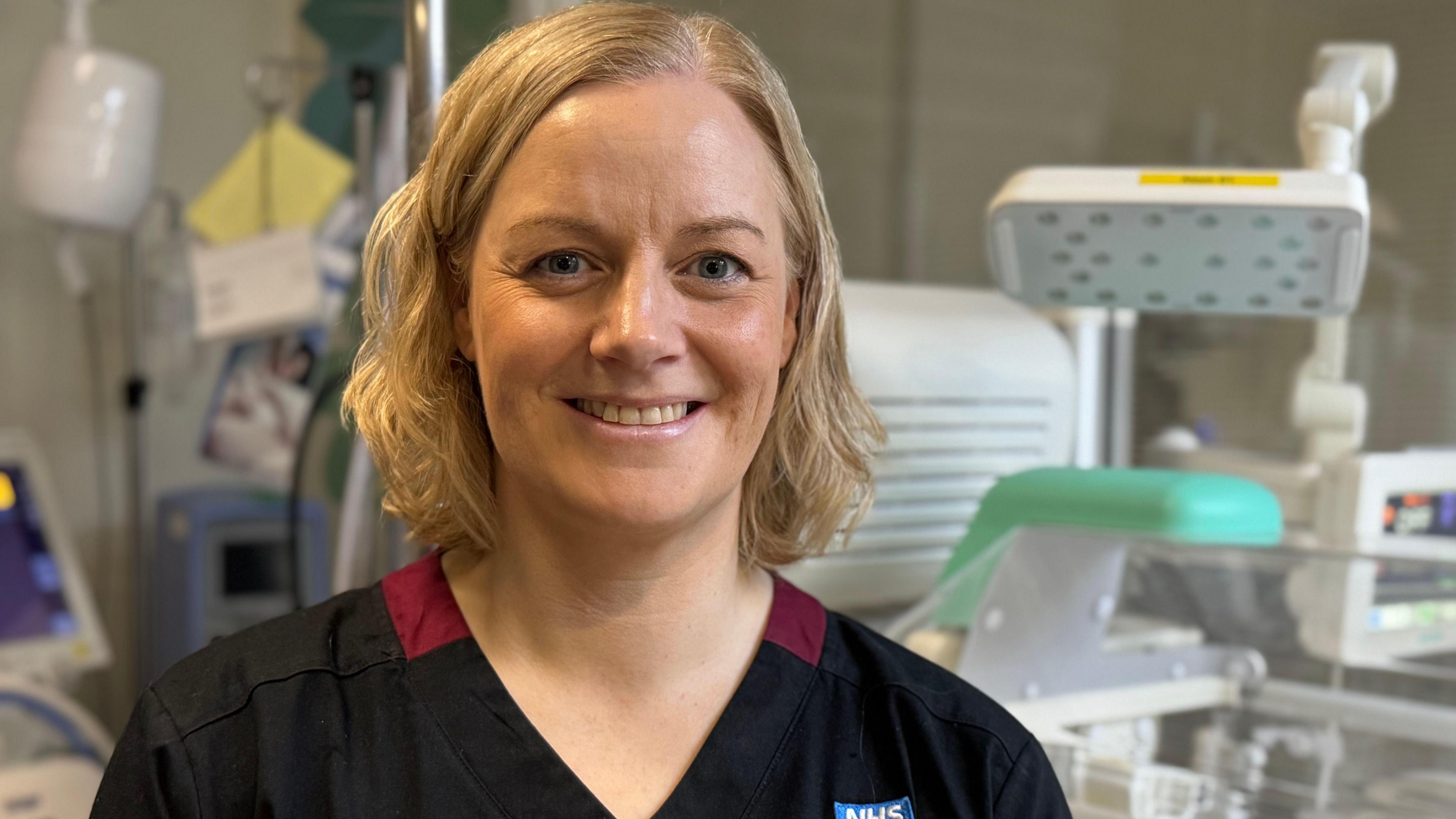 A woman with long blonde hair and wearing a black top with a small blue NHS badge, smiles while standing in front of various hospital machines and equipment.