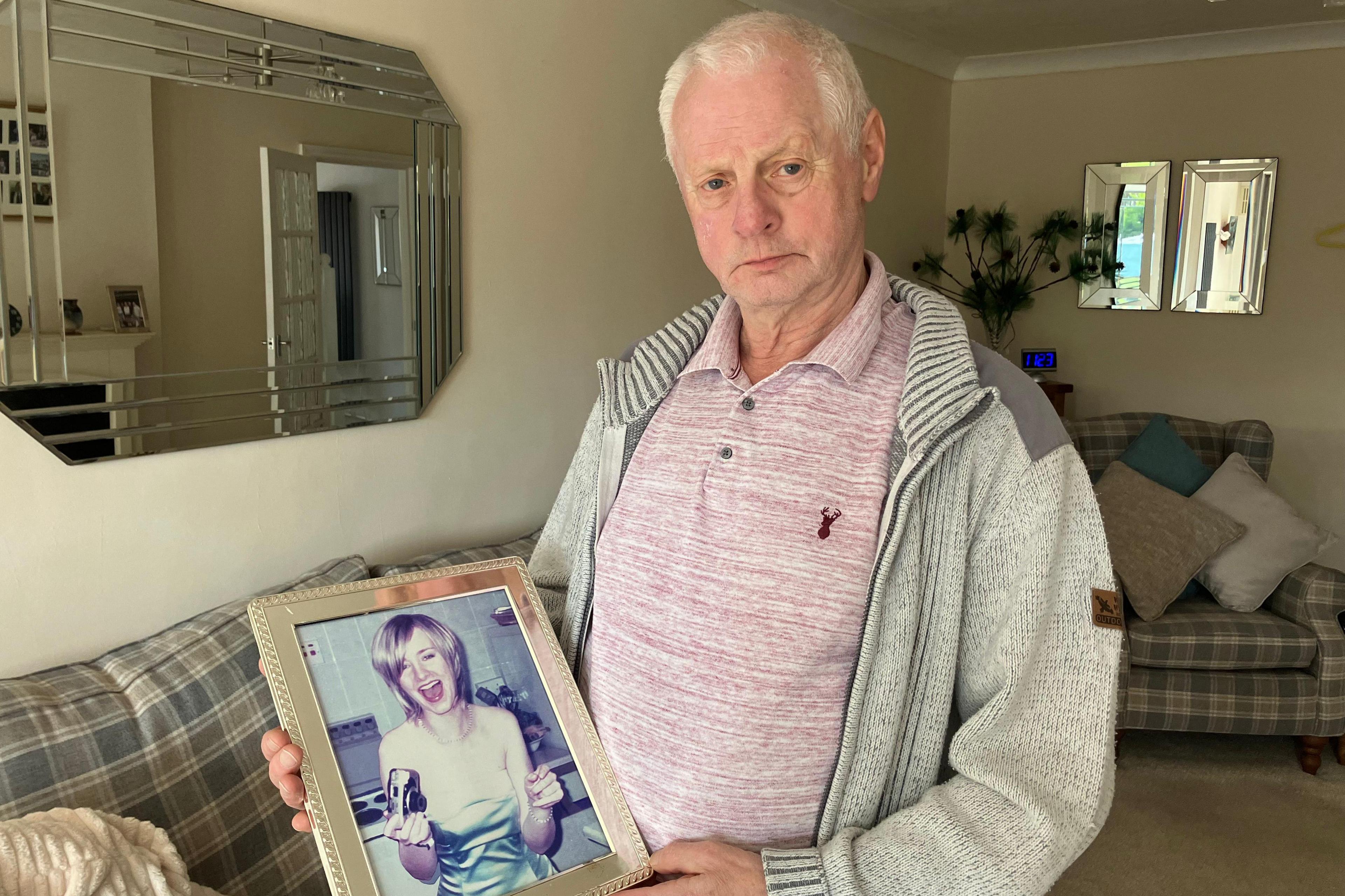 A picture of John Taylor holding a picture in a frame of his daughter Carrie inside his living room. He is wearing a pink polo shift and grey jumper.