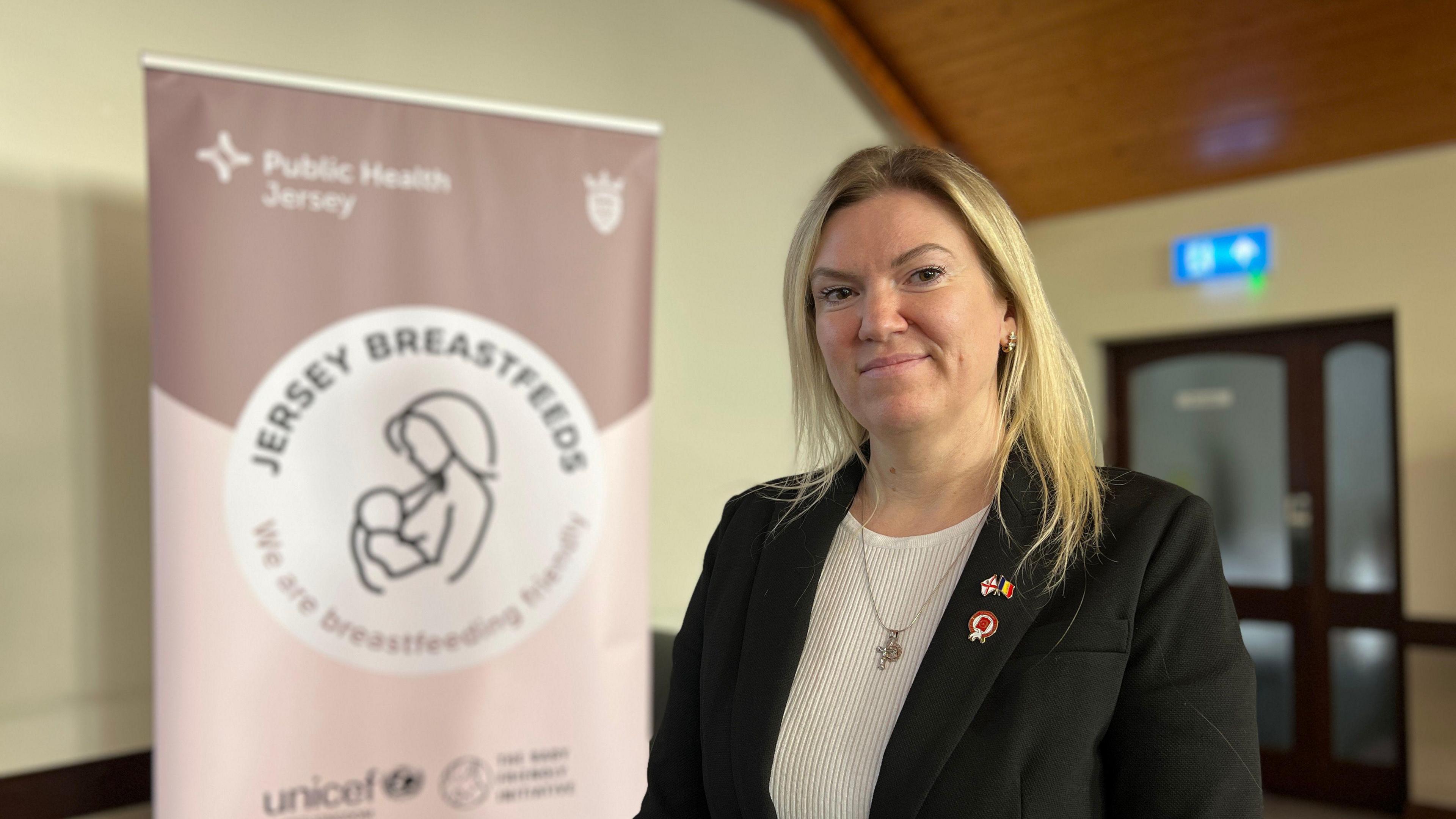 Raluca looks at the camera with a a sign behind her to her left which has the Breastfeeding Jersey Initiative logo on it. She is wearing a black jacket and has shoulder length blonde hair.