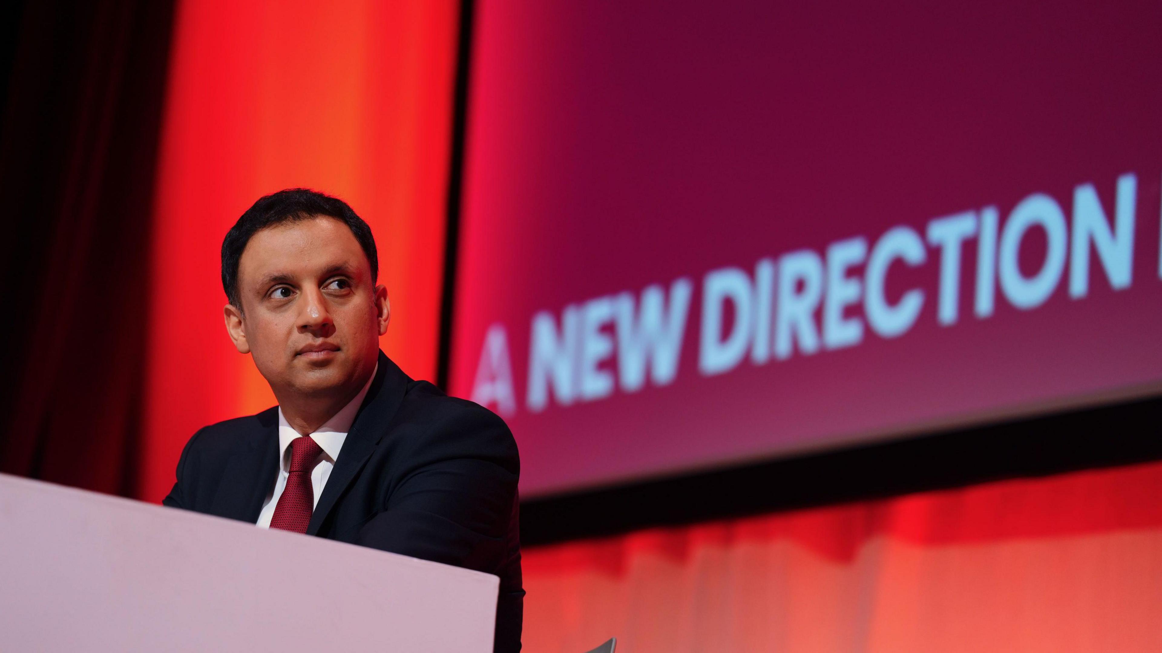 Anas Sarwar, a clean-shaven man with short black hair, a navy suit and red tie sits at a desk at a Labour conference, a red screen behind him displaying the words "a new direction".