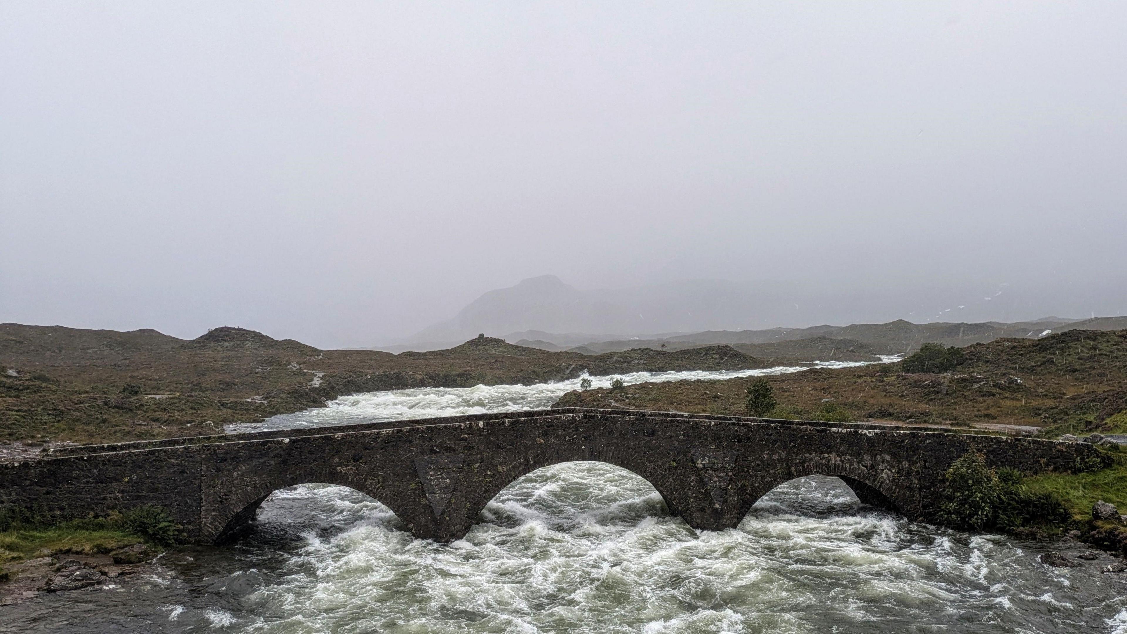 A very full, fast river close to the underside of an old stone bridge on Skye. It is very grey with low cloud and the surrounding hills cannot be seen for this.