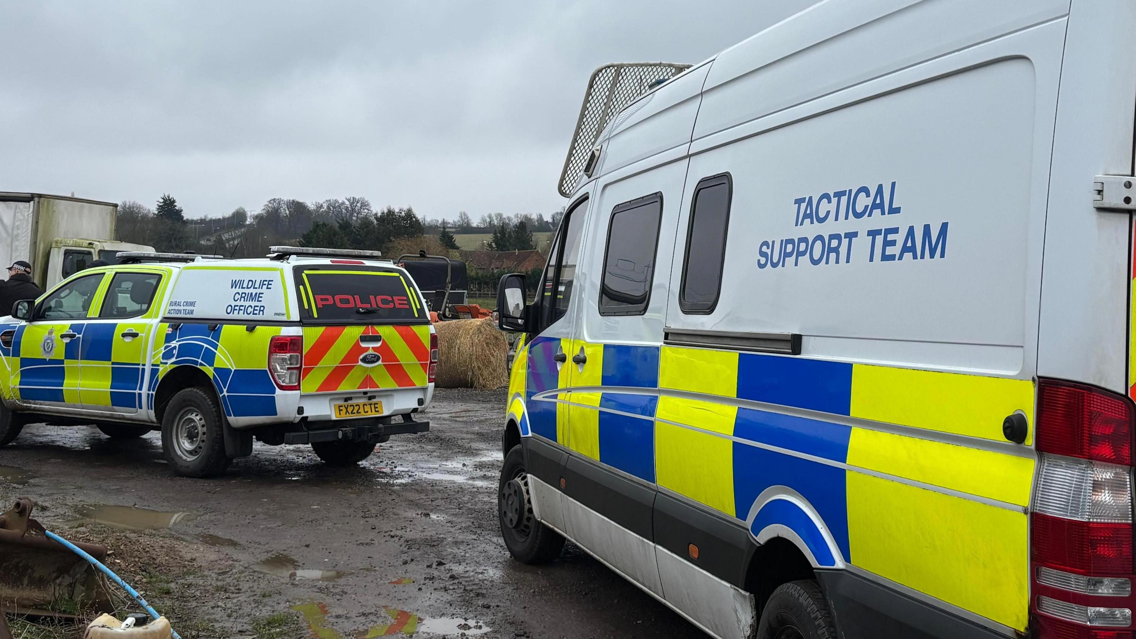 Two police tactical support vehicles are parked on what looks to be a farm. The ground is muddy with some puddles and there are fields and hay bales around them.