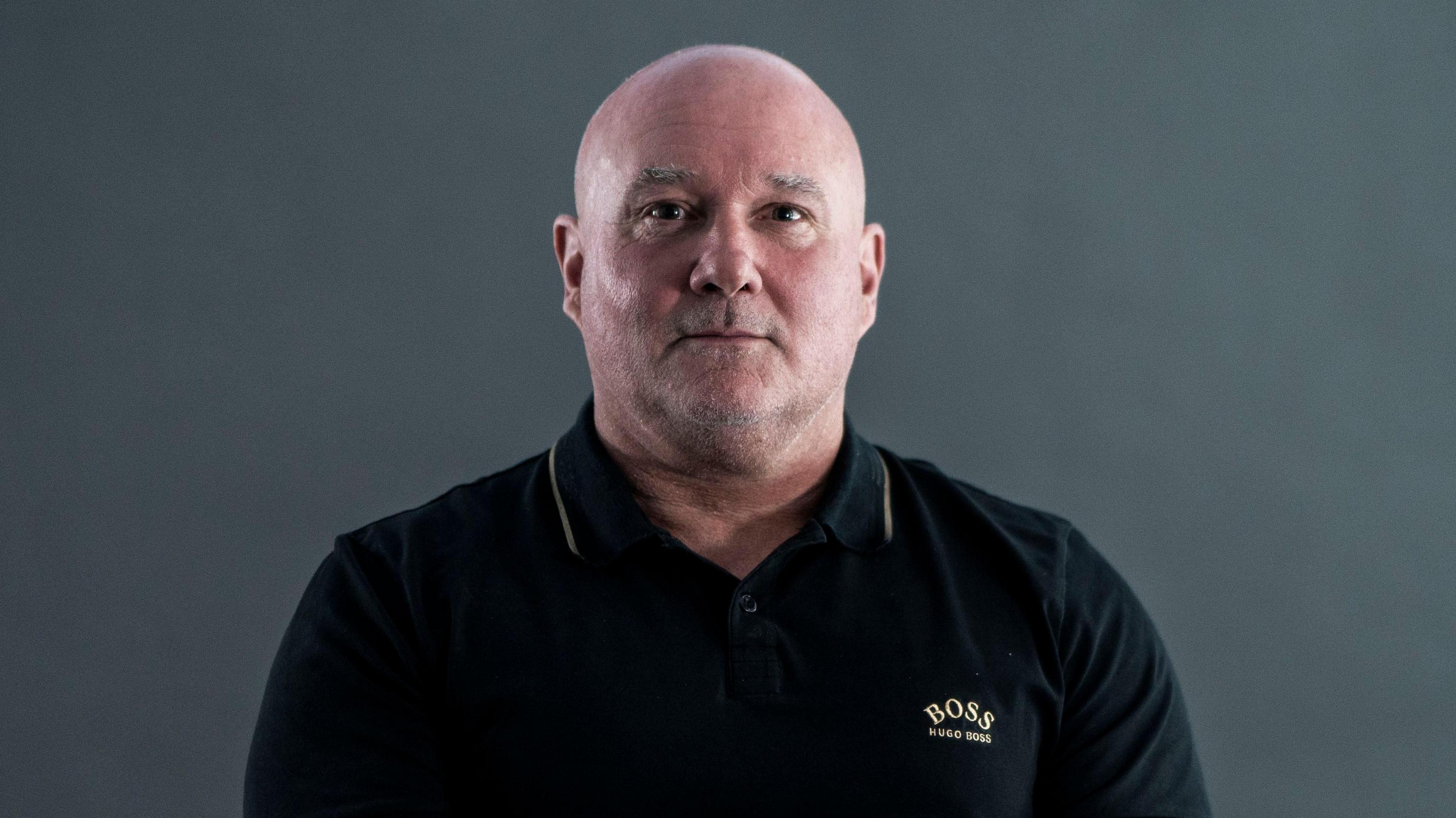Darren Attwood wearing a black polo shirt with gold detailing on it. He has a shaved head and looks serious.