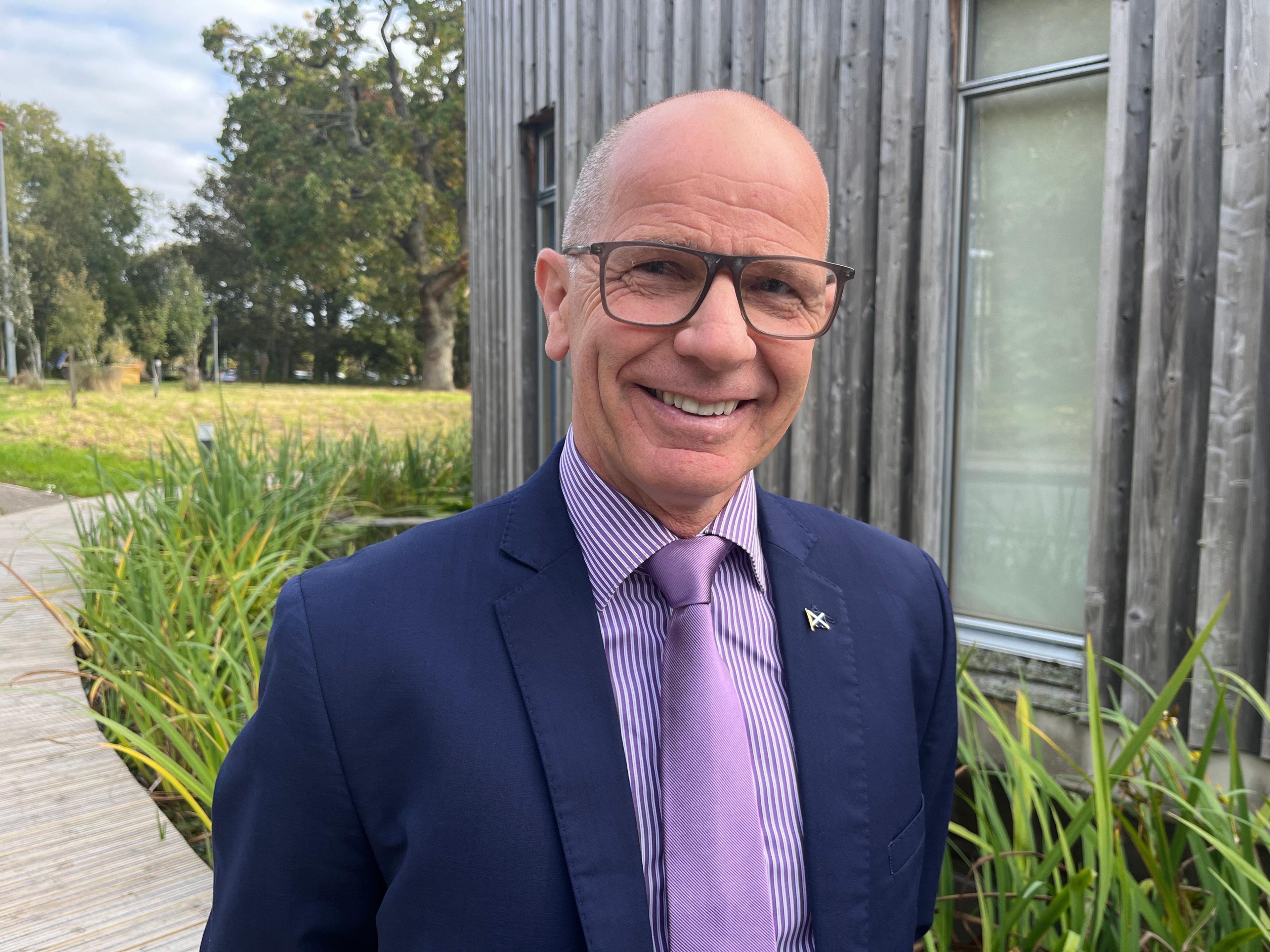 Jim Fairlie wearing a purple shirt and tie and a blue suit jackert, smiling to camera