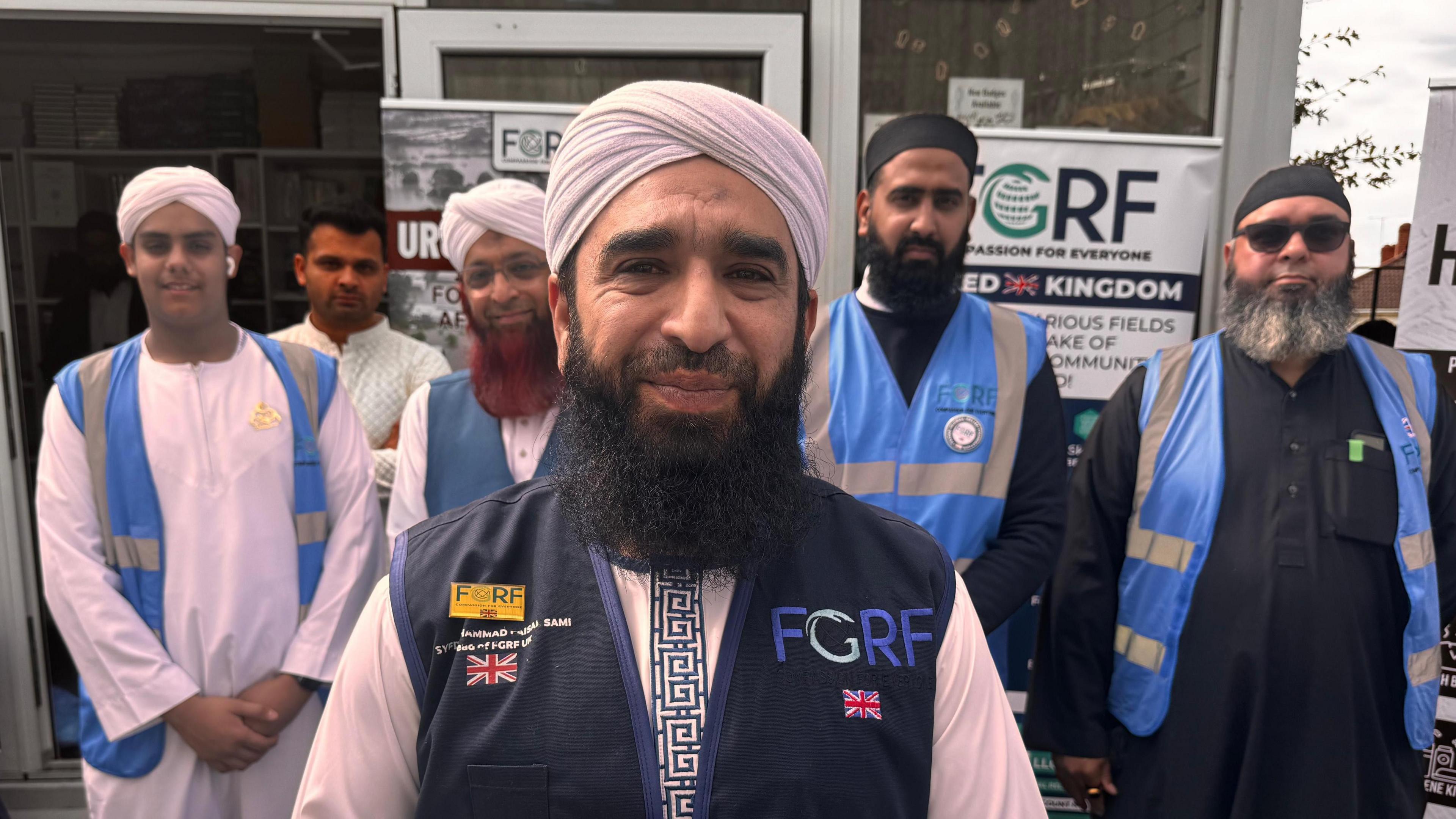 A group of six men, five of whom are wearing religious headwear, are posing for a photograph. They are standing in front of a building that has a glass door. Several of the men are wearing blue high-visibility jackets.