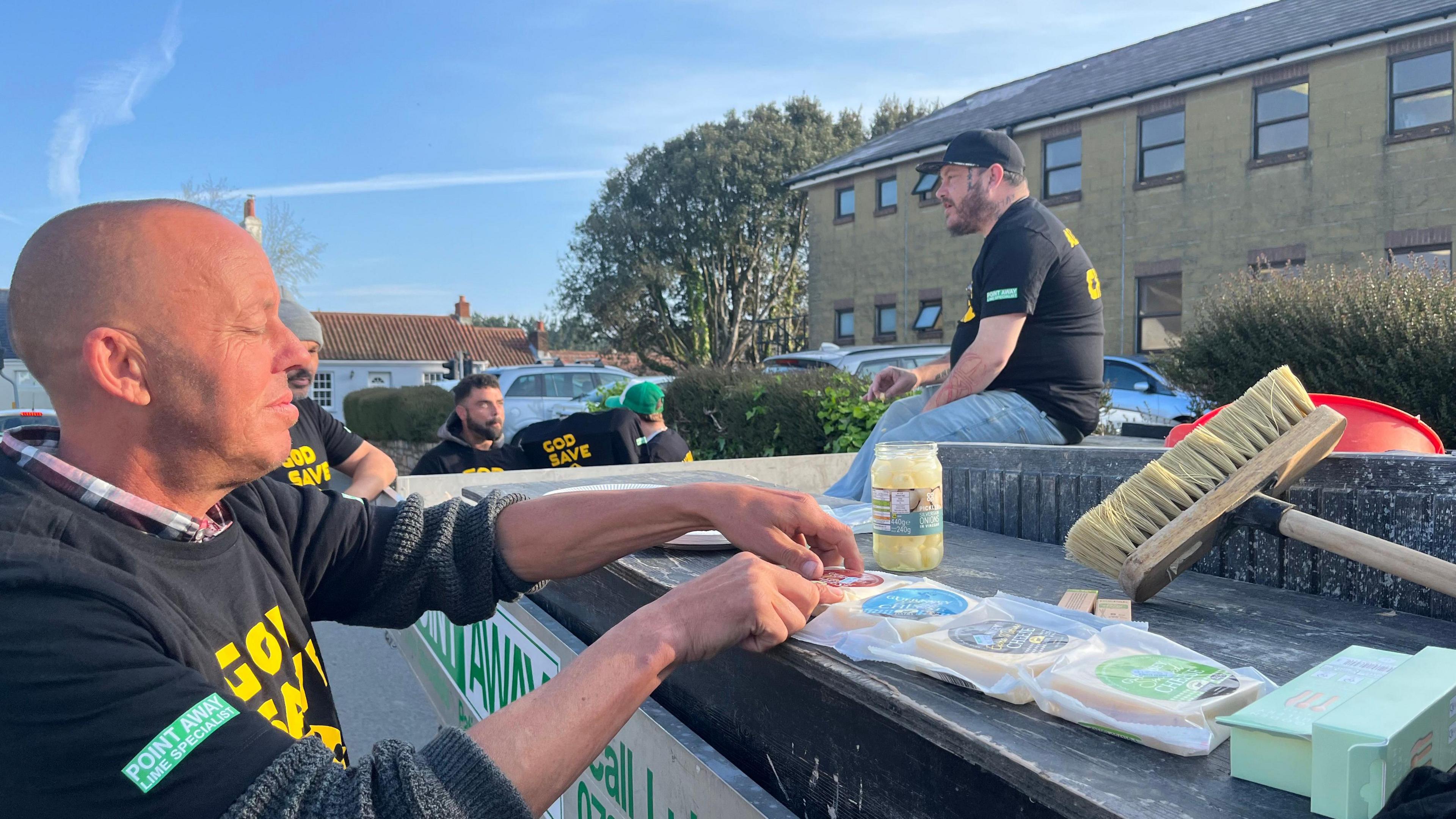 Five men stood around a van. There are four different types of cheese resting on the van and a broom and a jar of pickled onions. One man, waring a black t-shirt with yellow writing, is opening one of the packets of cheese. Another man, wearing a black and yellow t-shirt and blue jeans is sat on the van. The other three men are took behind the end of the van.