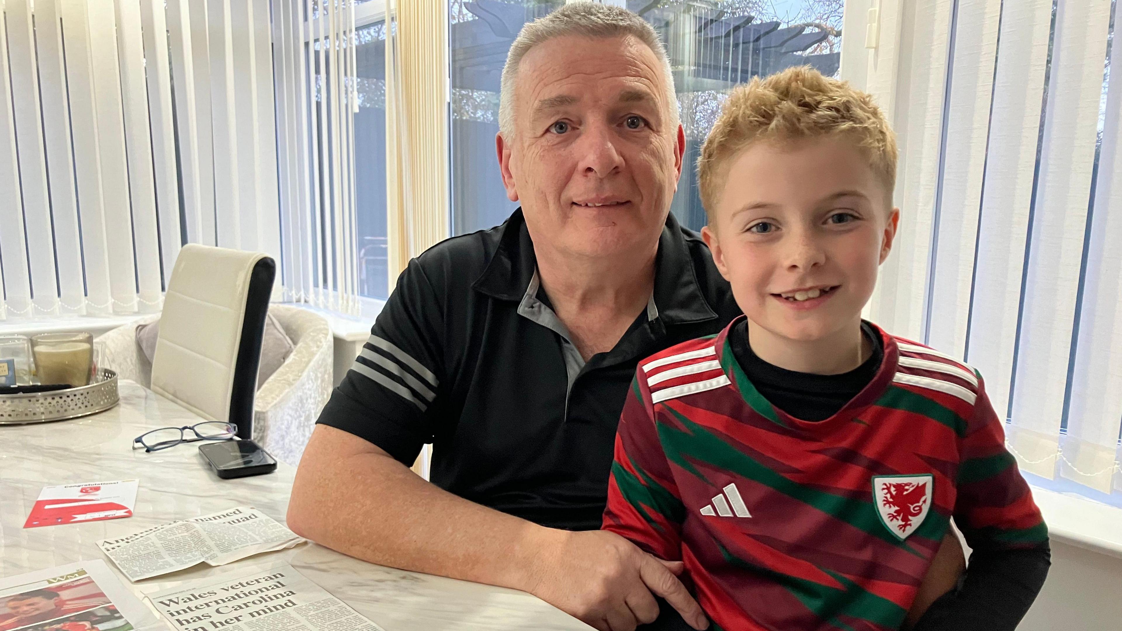 Angharad James' father sits with a child in a Wales football shirt at a table with cuttings which mention Angharad and the Wales team