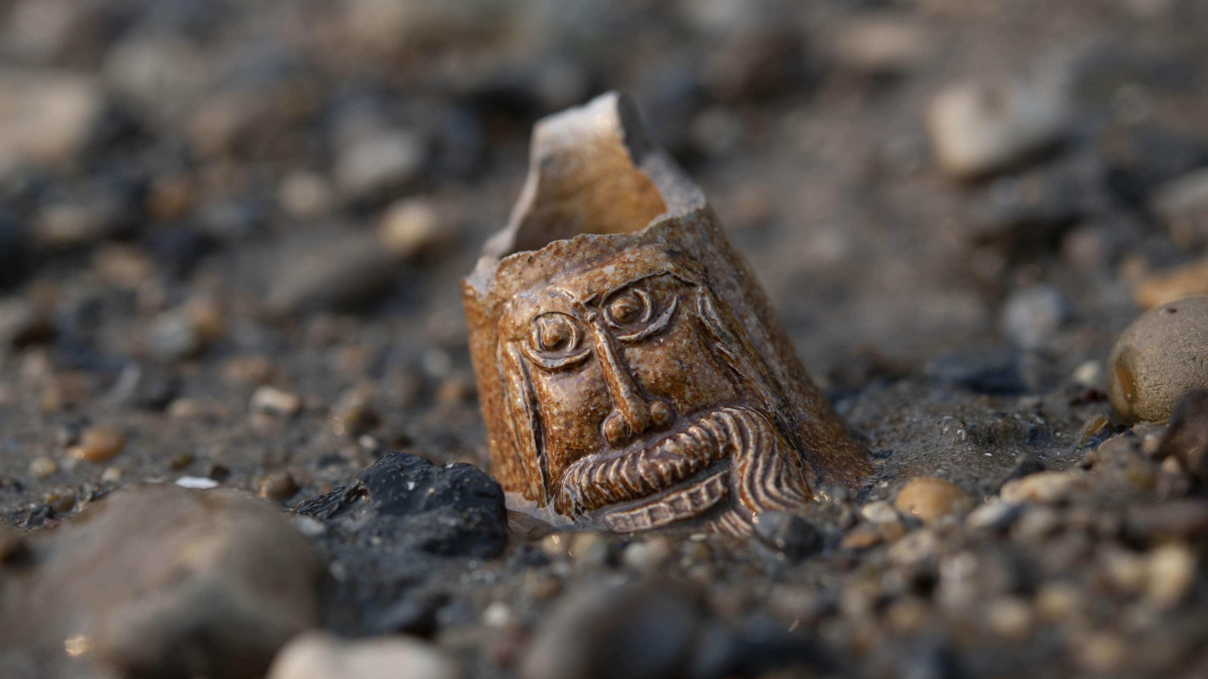 The neck from a stoneware bottle with a bearded face known as a Bartmann bottle from the 1500s to 1600s. The bearded face decorating the neck lies half-buried on the foreshore.