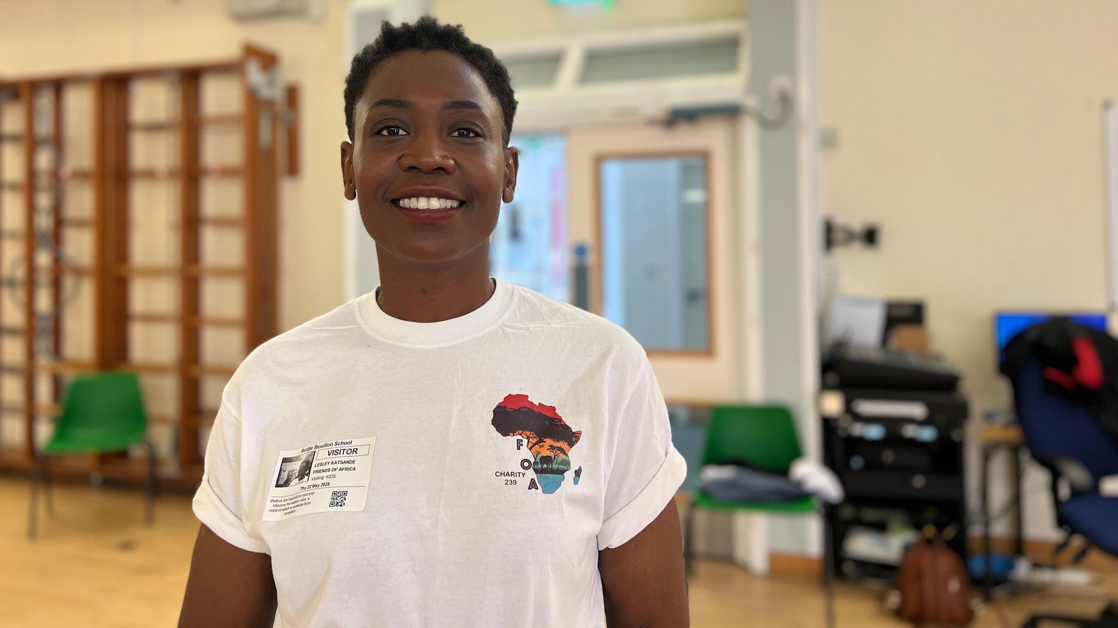 Lesley simles at the camera as she wears a white T-shirt with the charity logo on it. She has short black hair and is standing in a school's sports hall.