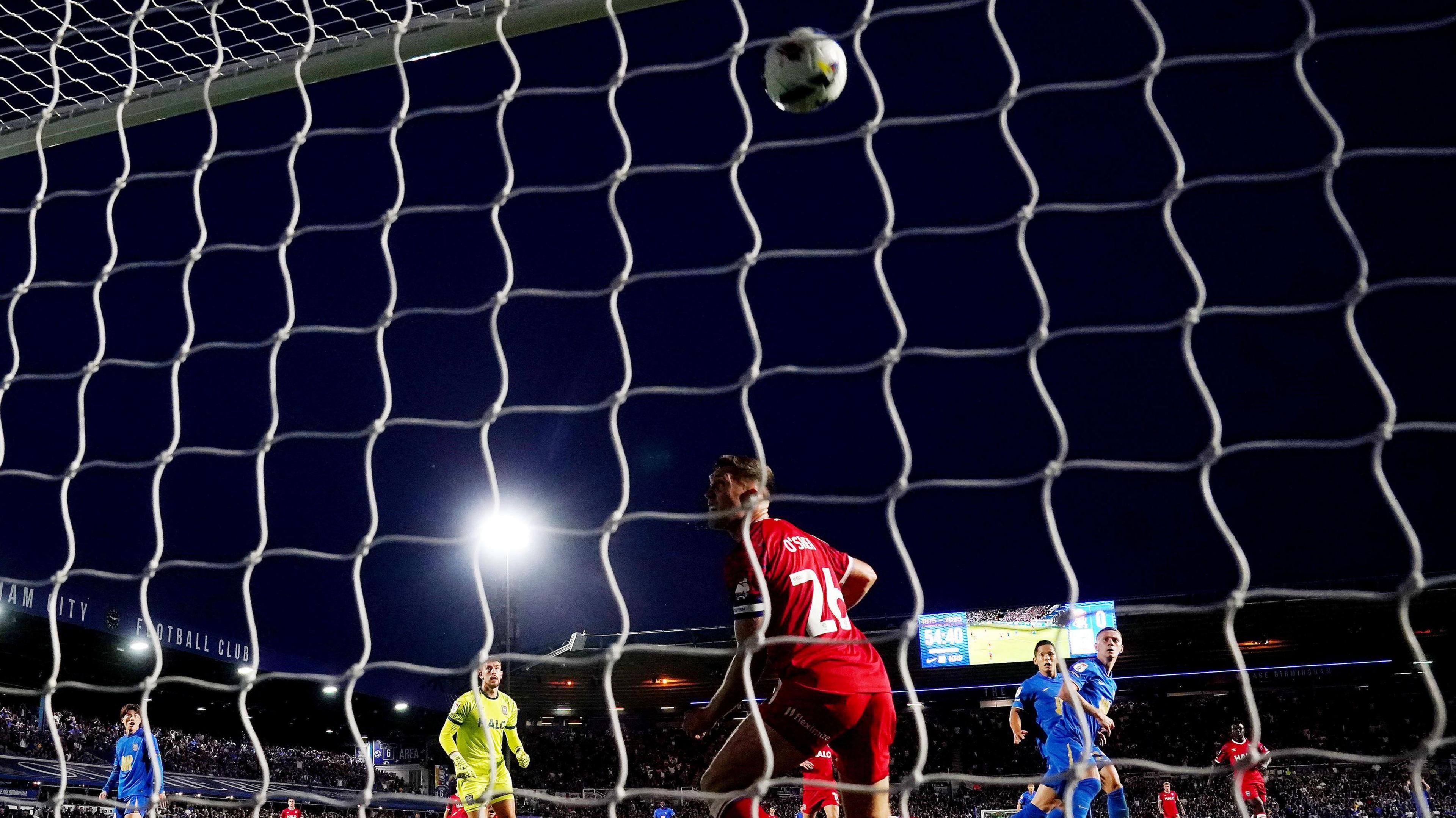A football match is taking place on a dark night. A ball is being kicked into the net and players are watching it in the air. We can see a floodlight shining bright and fans in the background. There is also a bright screen showing the score and the time. 