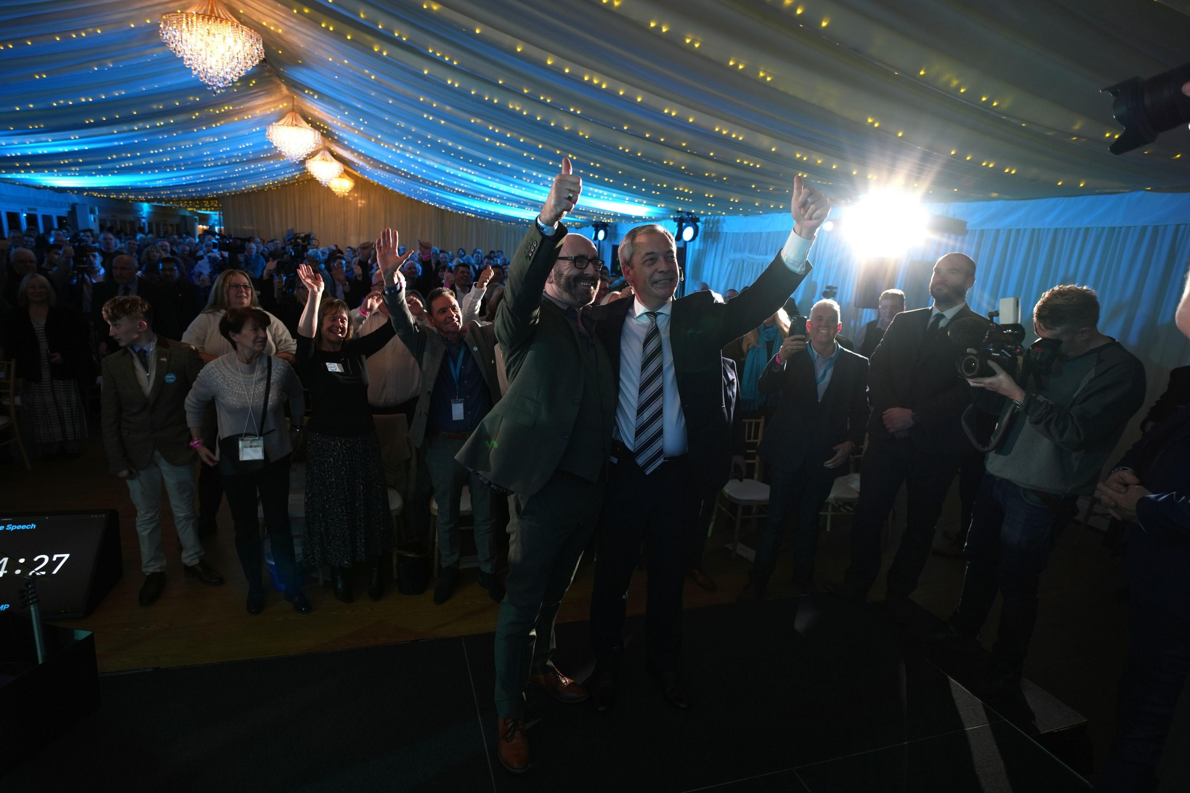 Farage has his arm around another man's shoulder and they make a 'thumbs-up' gesture in the test. Cheering suppoters can be seen in the background