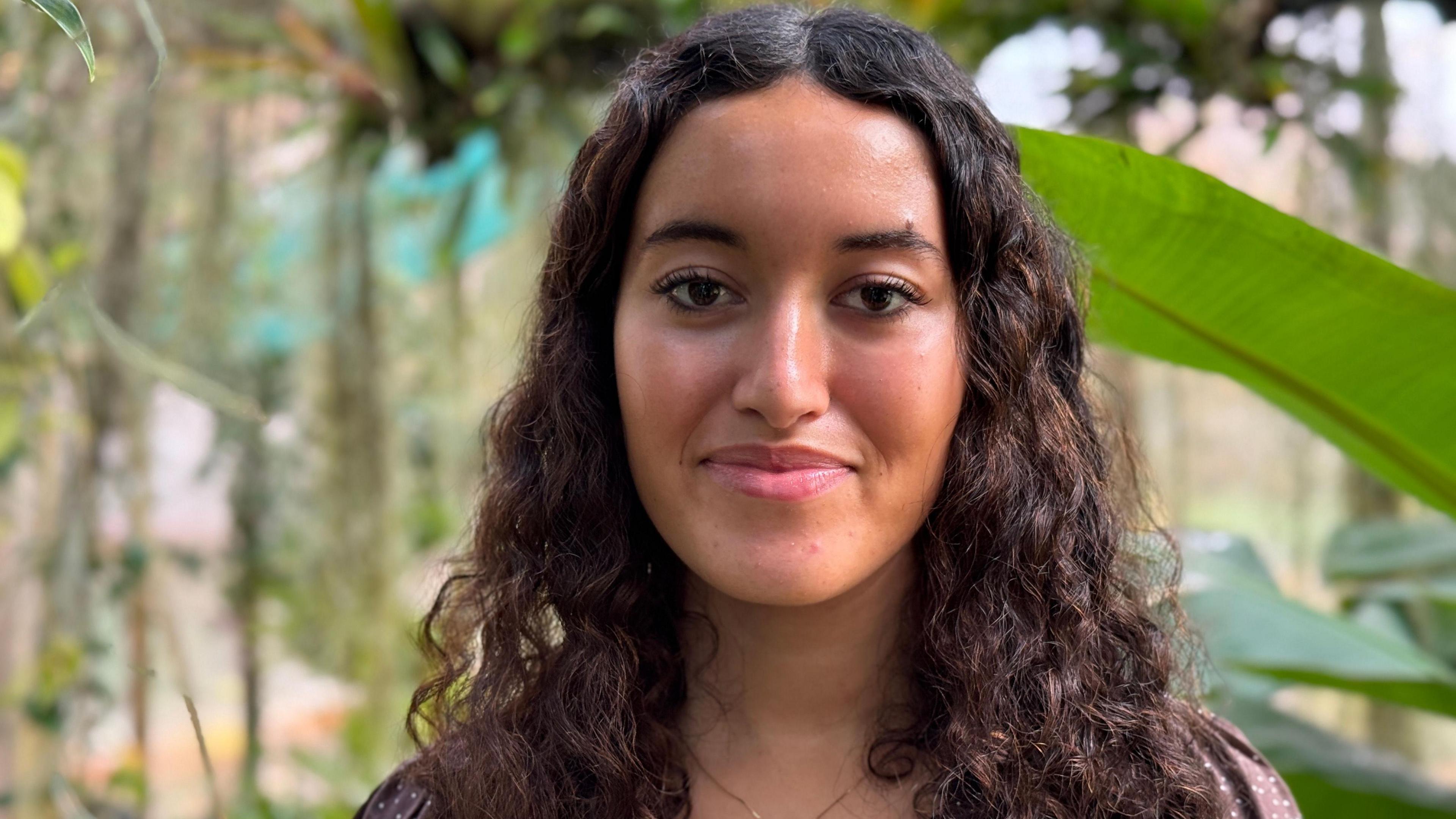 A girl with long, curly brown hair is smiling at the camera. Behind her there are green leaves and brown vines