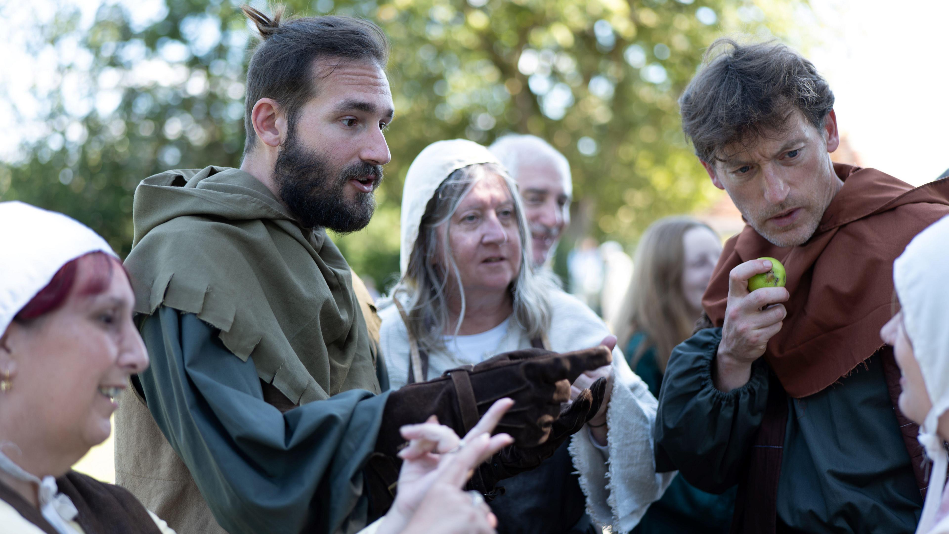 Six actors during a dress rehearsal, wearing medieval costumes, with one man eating an apple