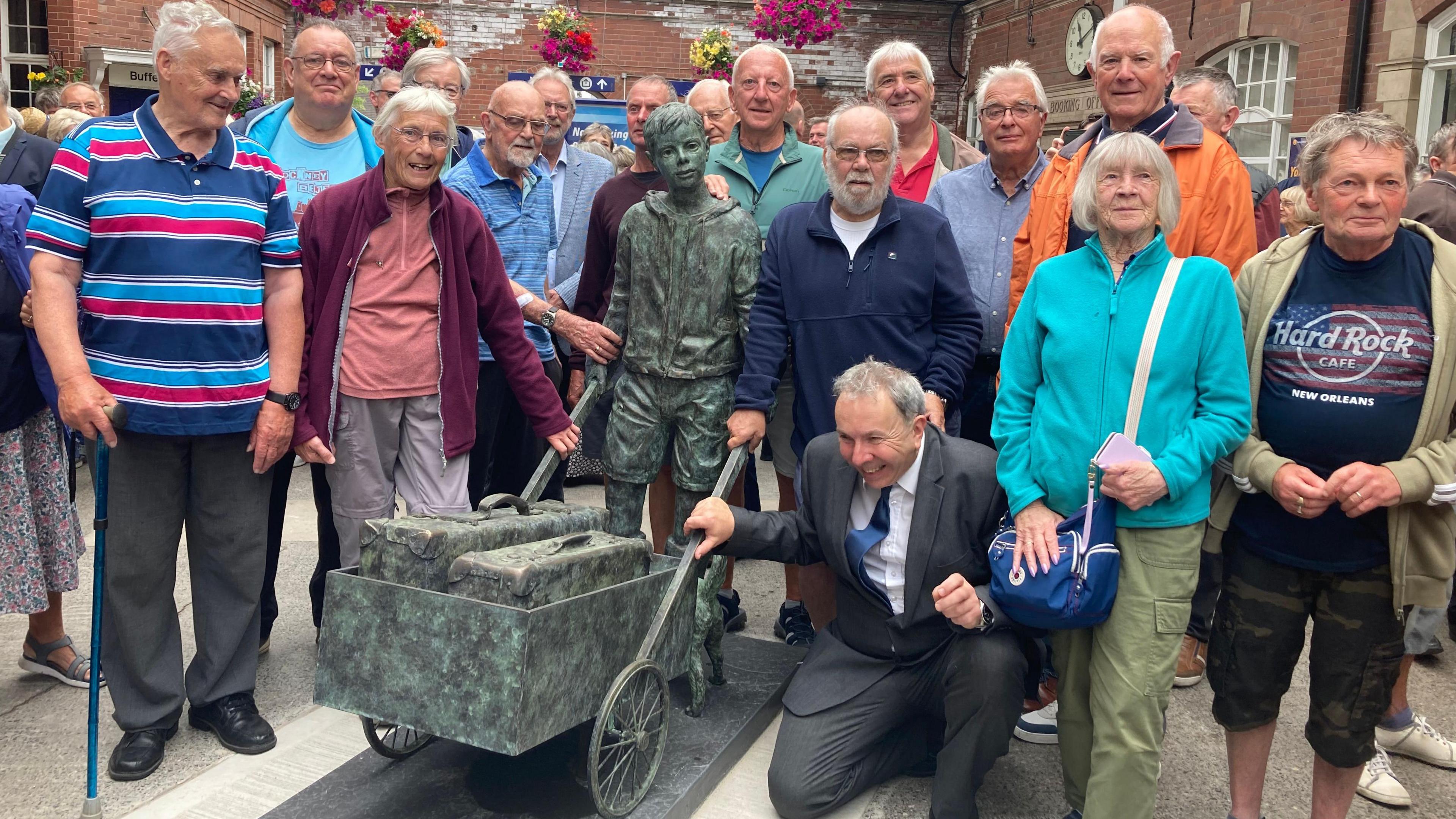 A crowd of about 15 people, mainly elderly men and women, gathers around a grey-green sculpture of a boy pushing a barrow at Bridlington railway station