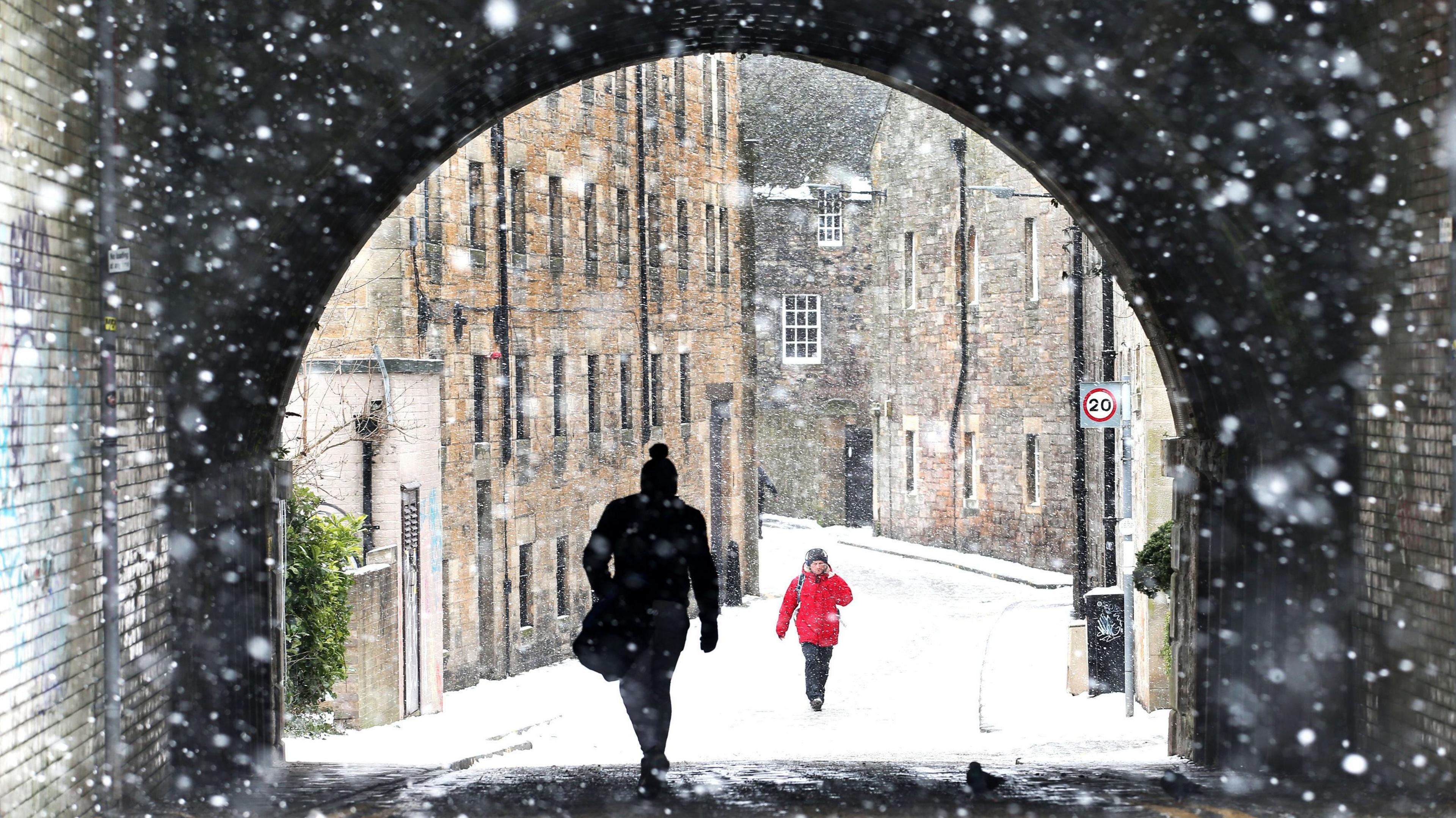 A view through falling snow down a narrow snow-covered road framed by a brick arch