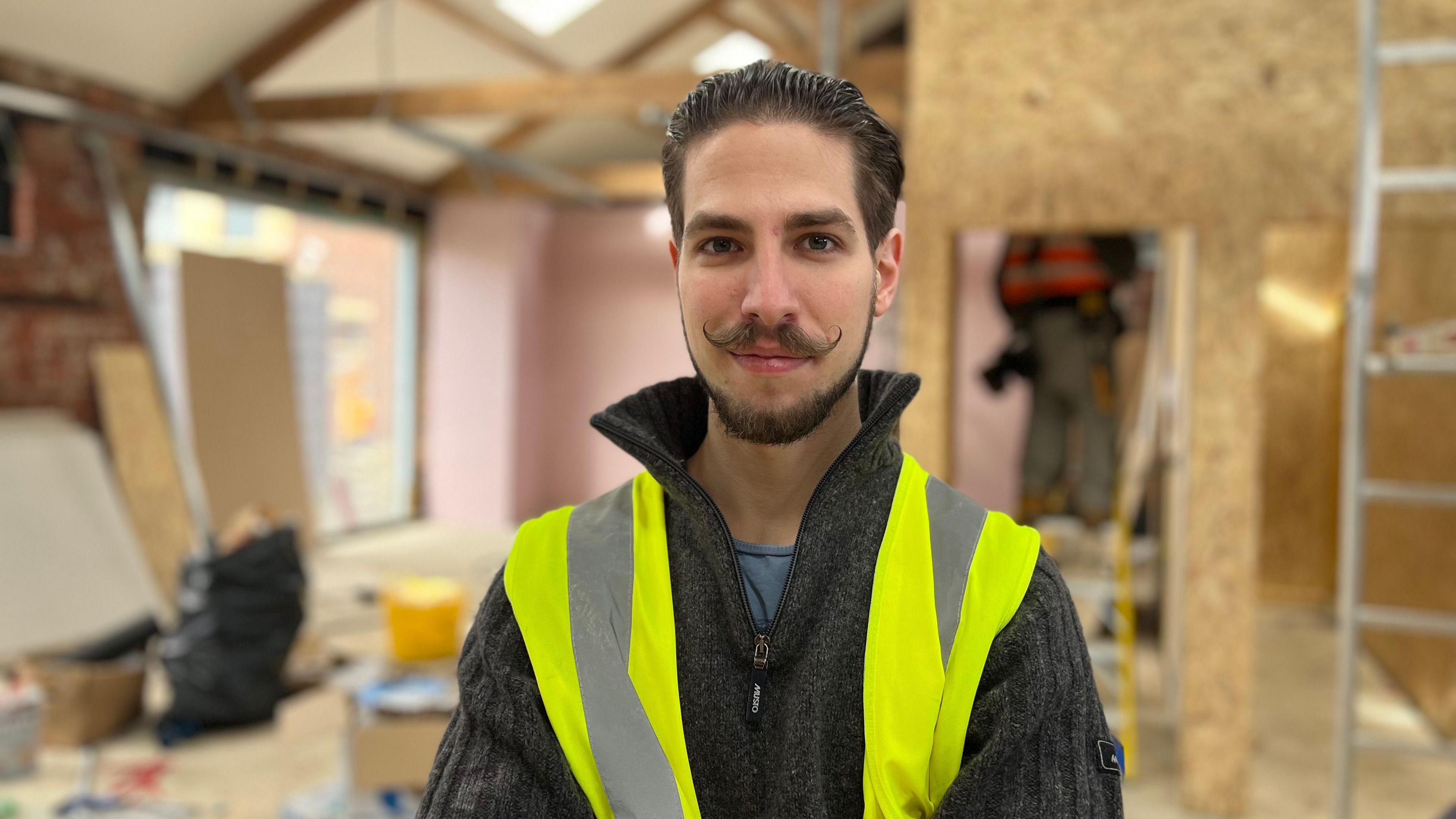 A picture of a man with high vis jacket in building site- he has a unusual moustache