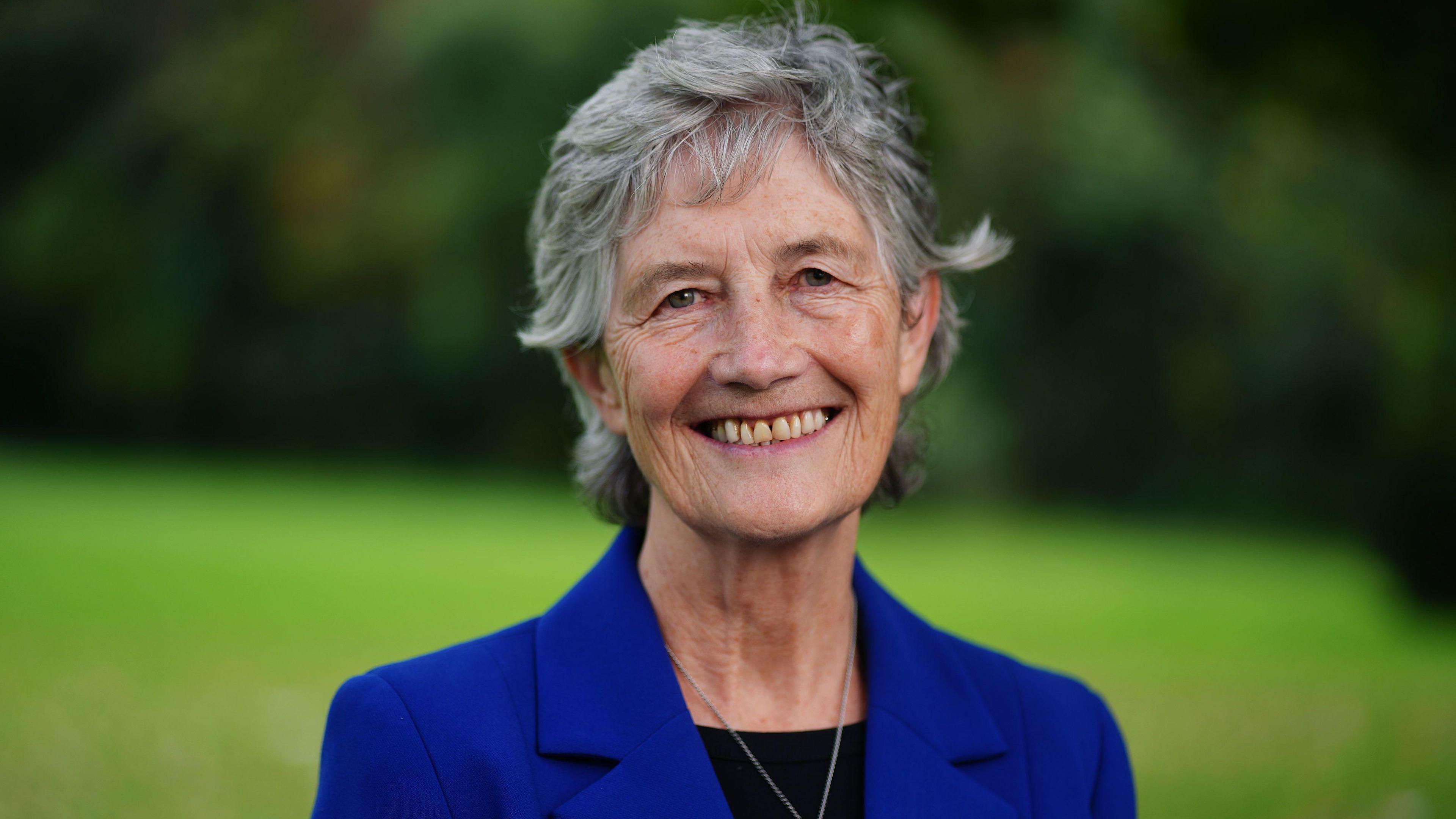 Catherine Connolly - a woman with short, grey hair - looking directly at the camera and smiling broadly during a photoshoot.  She is wearing a royal blue suit jacket over a black crew neck top with a silver pendant necklace.  She is standing in a lawned area but the background is out of focus.