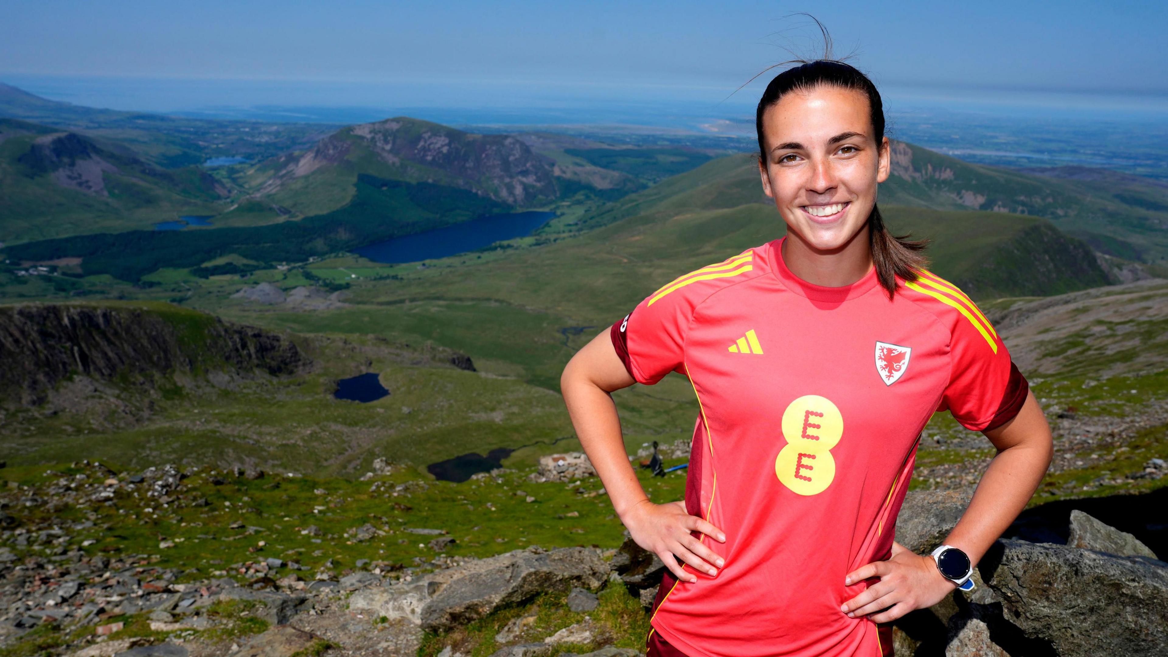 Olivia Clark is smiling at the camera. She is wearing a red football shirt, which has the Welsh dragon emblem to the right. Her hands are on her hips, and her brown hair is pulled back into a ponytail. She is standing on top of a mountain.