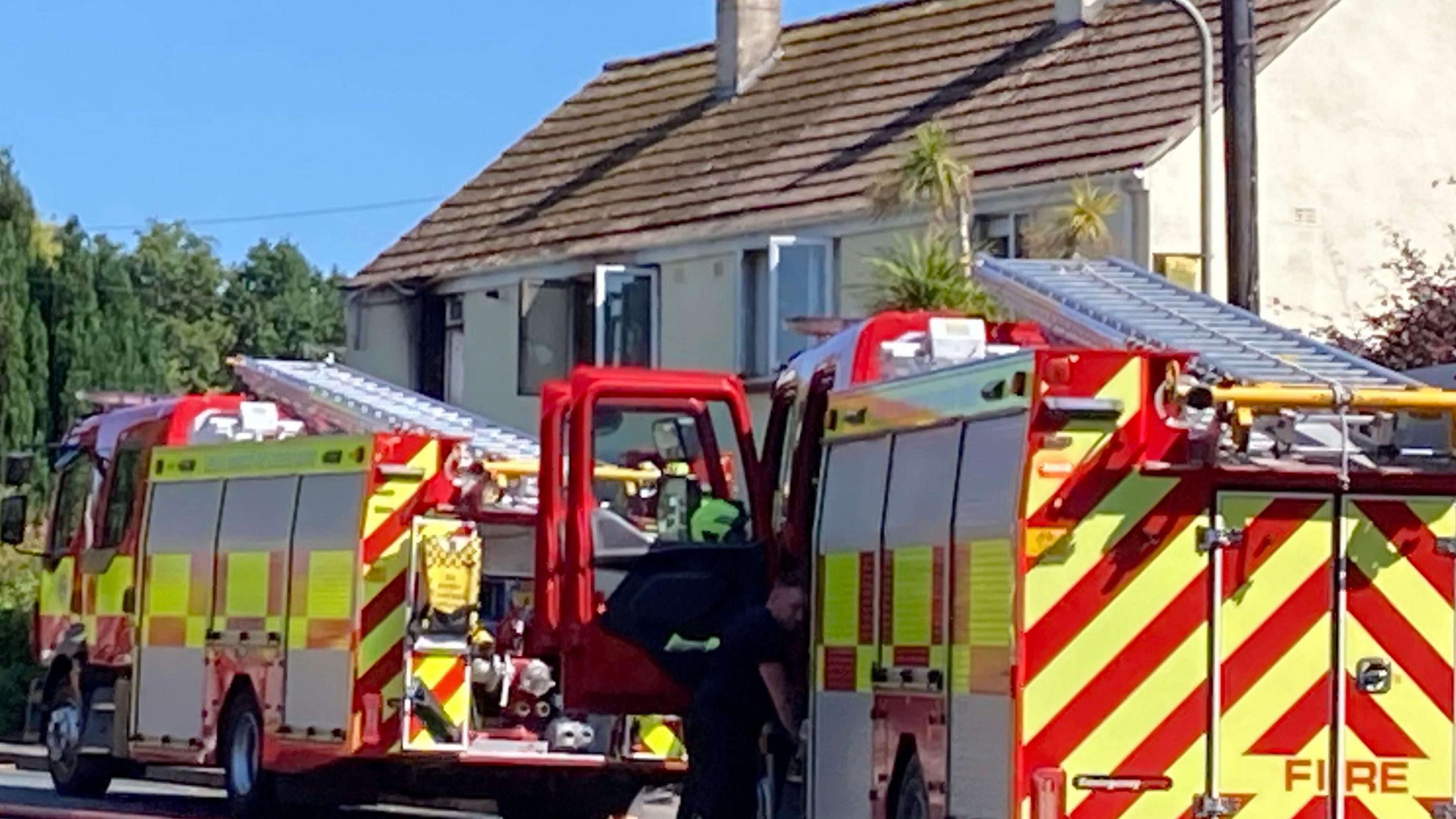 Two fire engines outside a house with signs of smoke at a window.