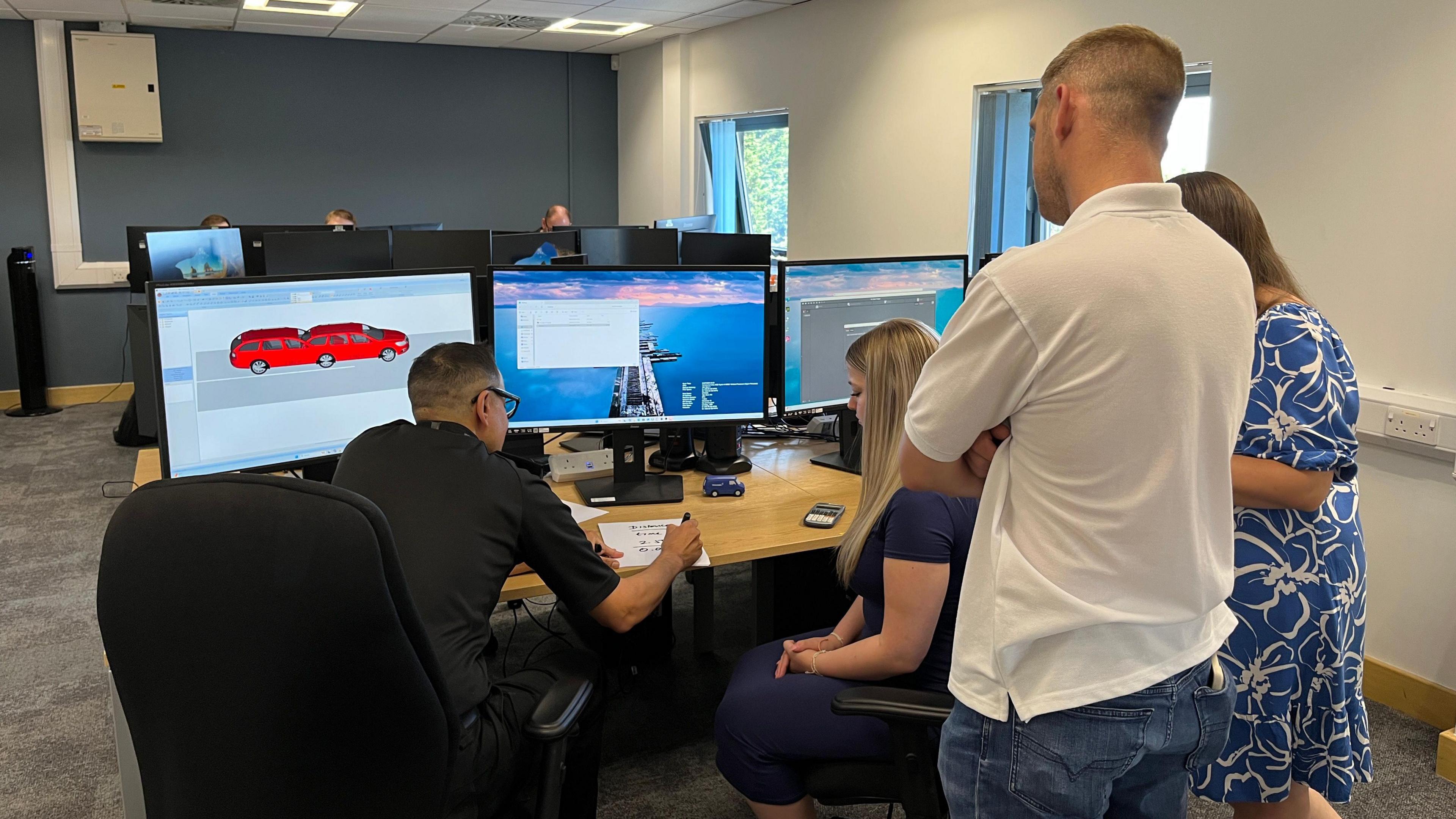 A teenage girl with long blonde hair and a navy top is sitting down at a desk of computers, next to a police officer who has glasses and a black shirt. On the screen in front of him are diagrams of two red cars. A man and a woman stand above them