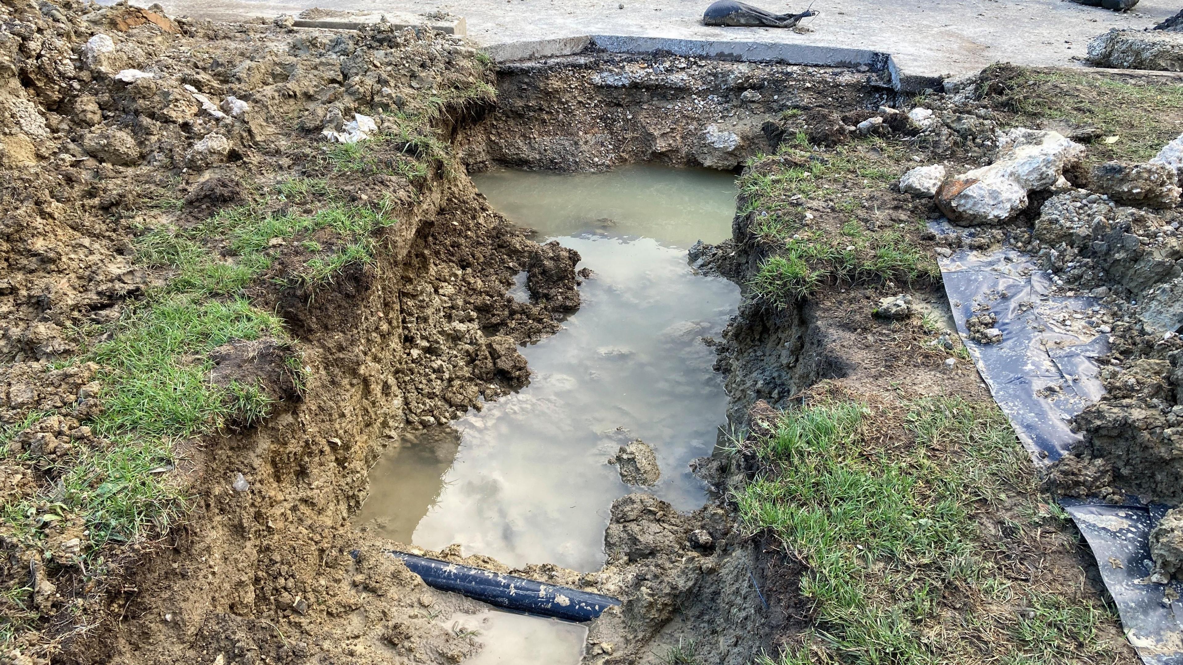 A trench dug in the grass reveals waterlogged ground with a single exposed black pipe.