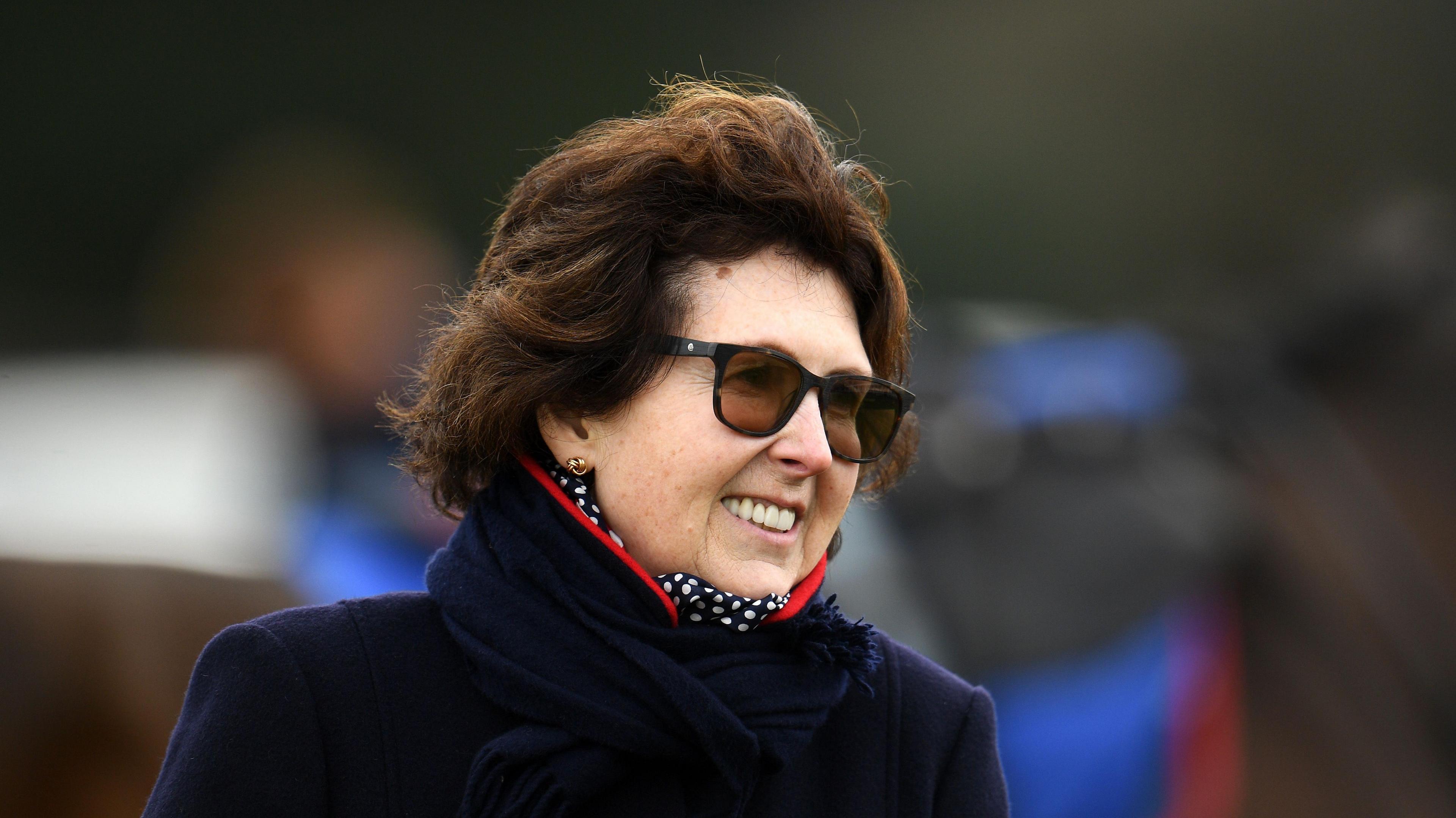 A woman with shoulder length wavy brown hair is pictured wearing brown sunglasses and a navy blue coat with a navy blue and white dotted neck scarf. 