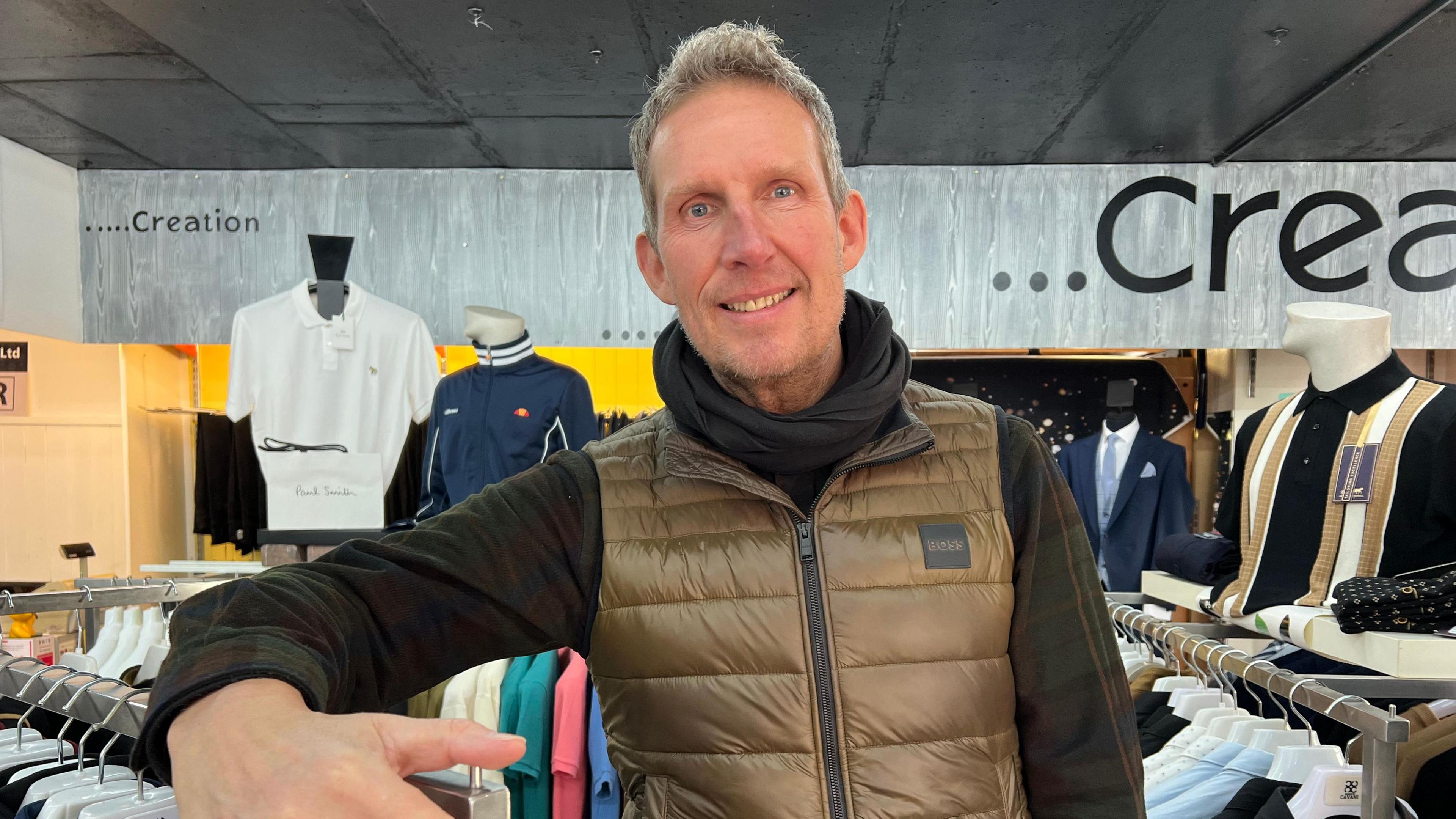 A man stands among racks of clothes in his store. He is tall with quiffed hair and wears a gold gillet, checked shirt and black snood.