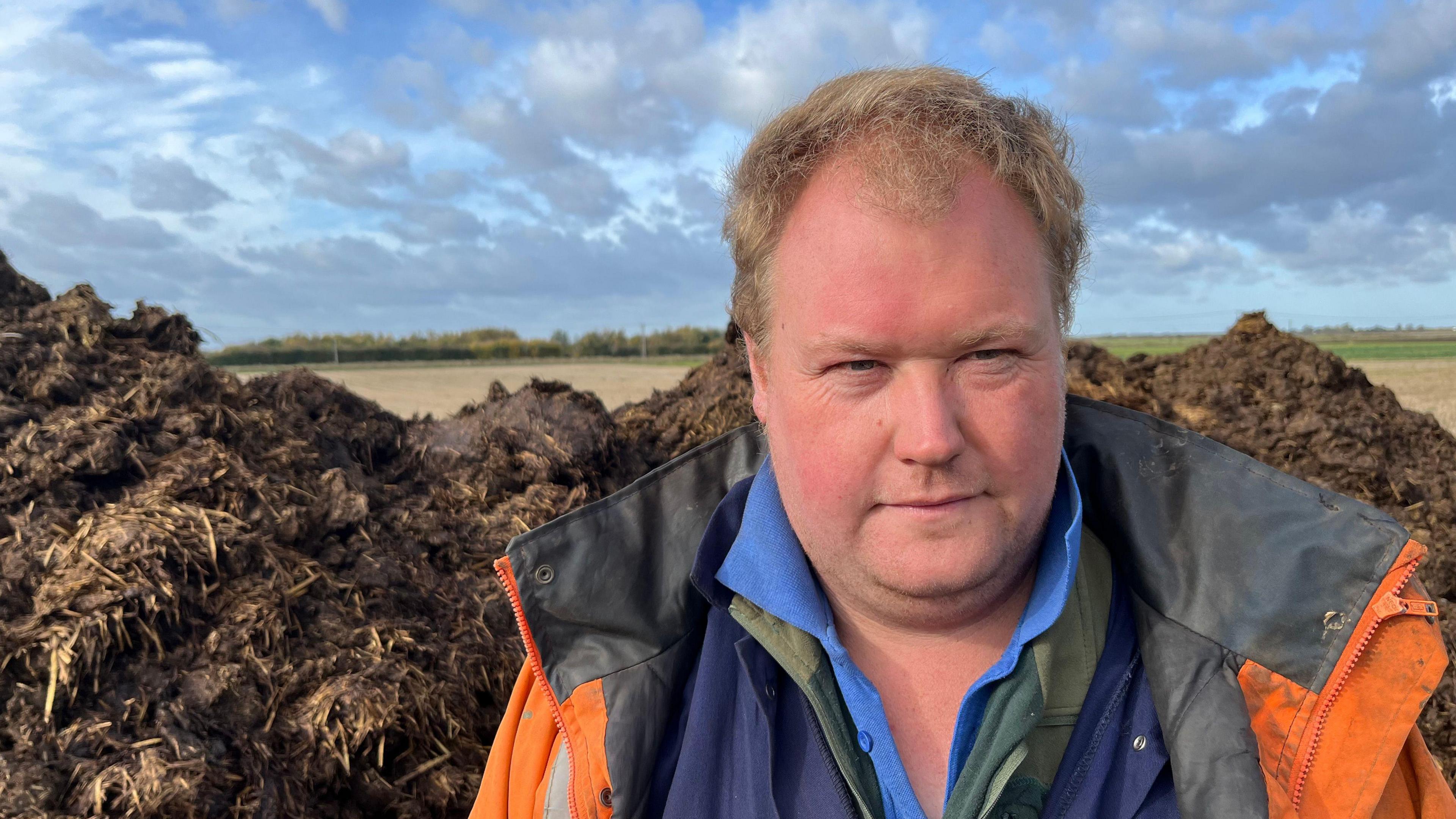 Richard Freeman has short red hair and is wearing a high-visibility jacket over a blue fleece over a green fleece over a lighter blue shirt.
Behind him, in a farm field is a large heap of manure.