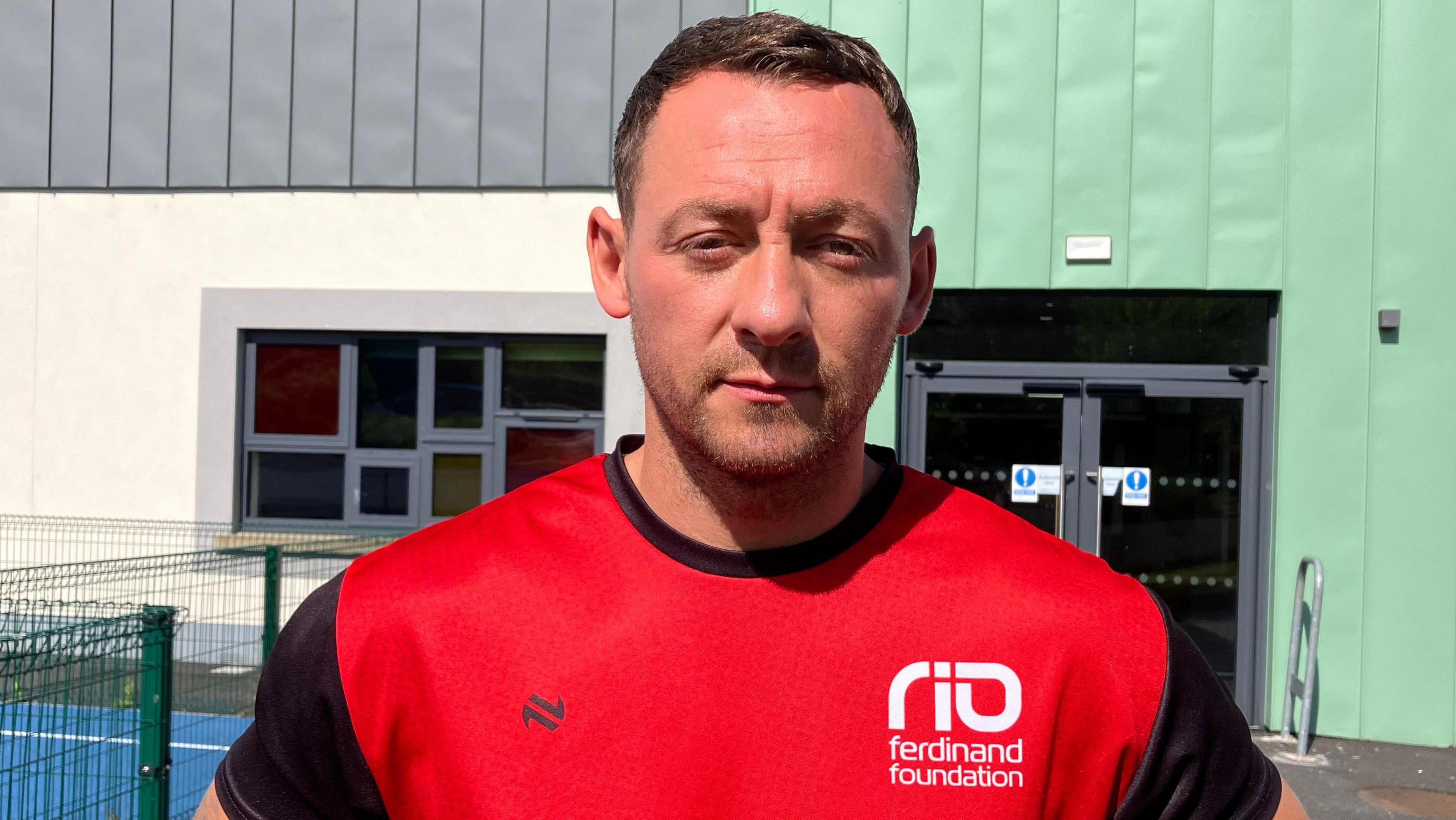 A man in a red t-shirt with black sleeves. He is standing in front of a blue sports' pitch.