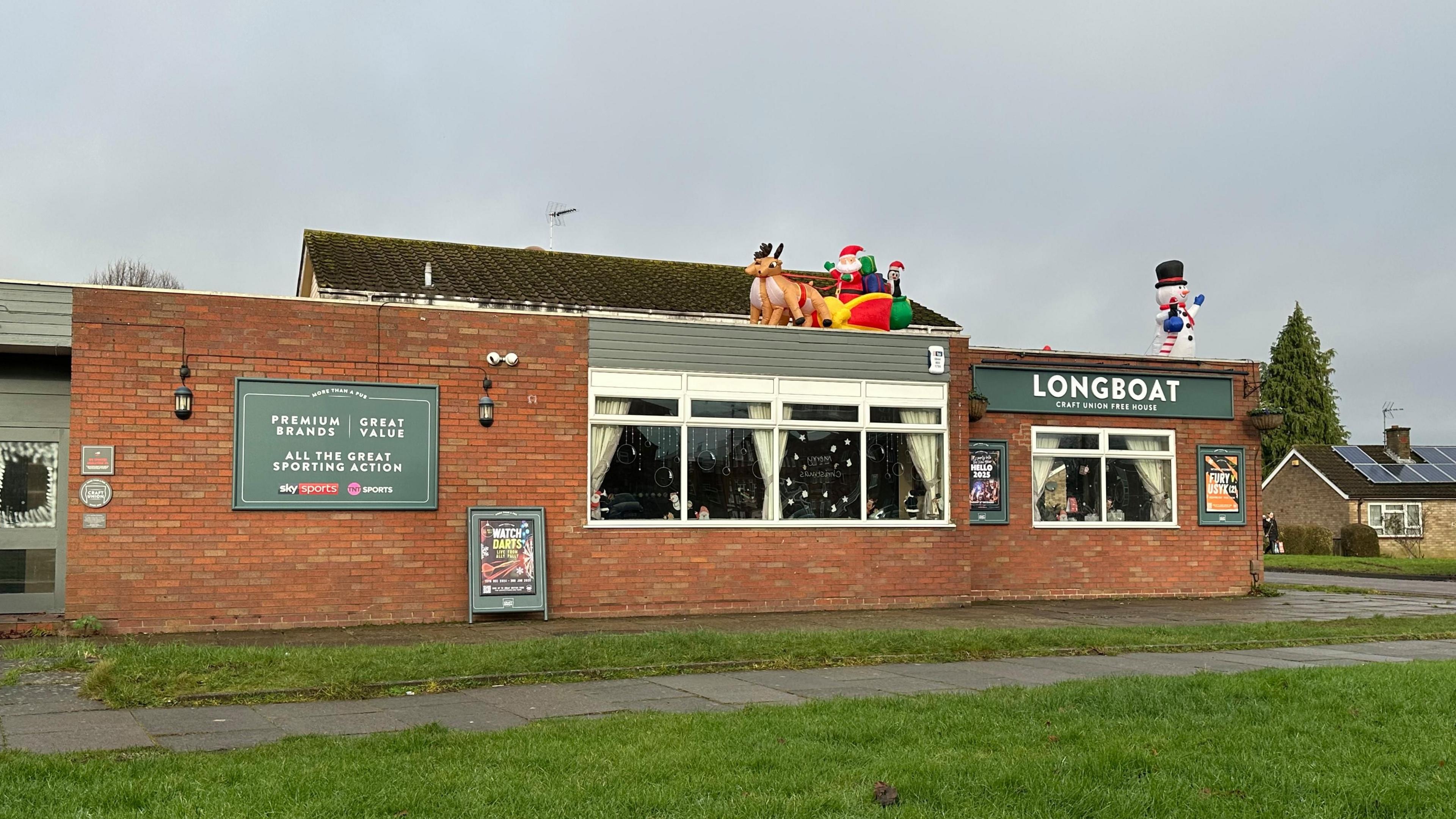 Red brick one-storey pub building with green signage, with windows in the centre and the right. Green grass in front of the building with path in between.