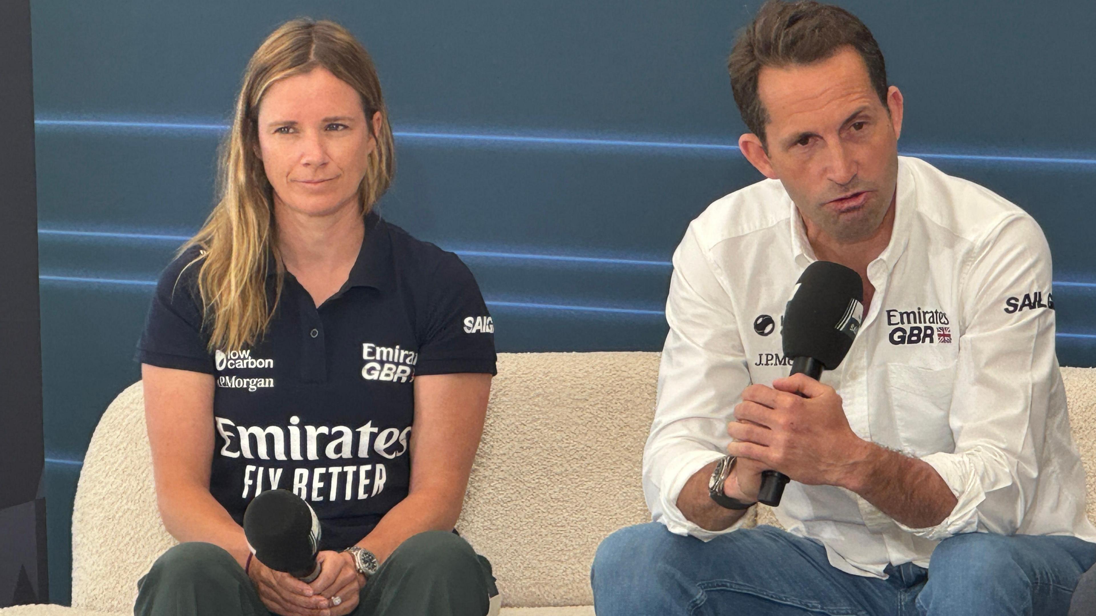 Hannah Mills wearing a blue GBR t-shirt and holding a microphone sat next to Sir Ben Ainslie who is wearing a white GBR shirt and talking into a microphone