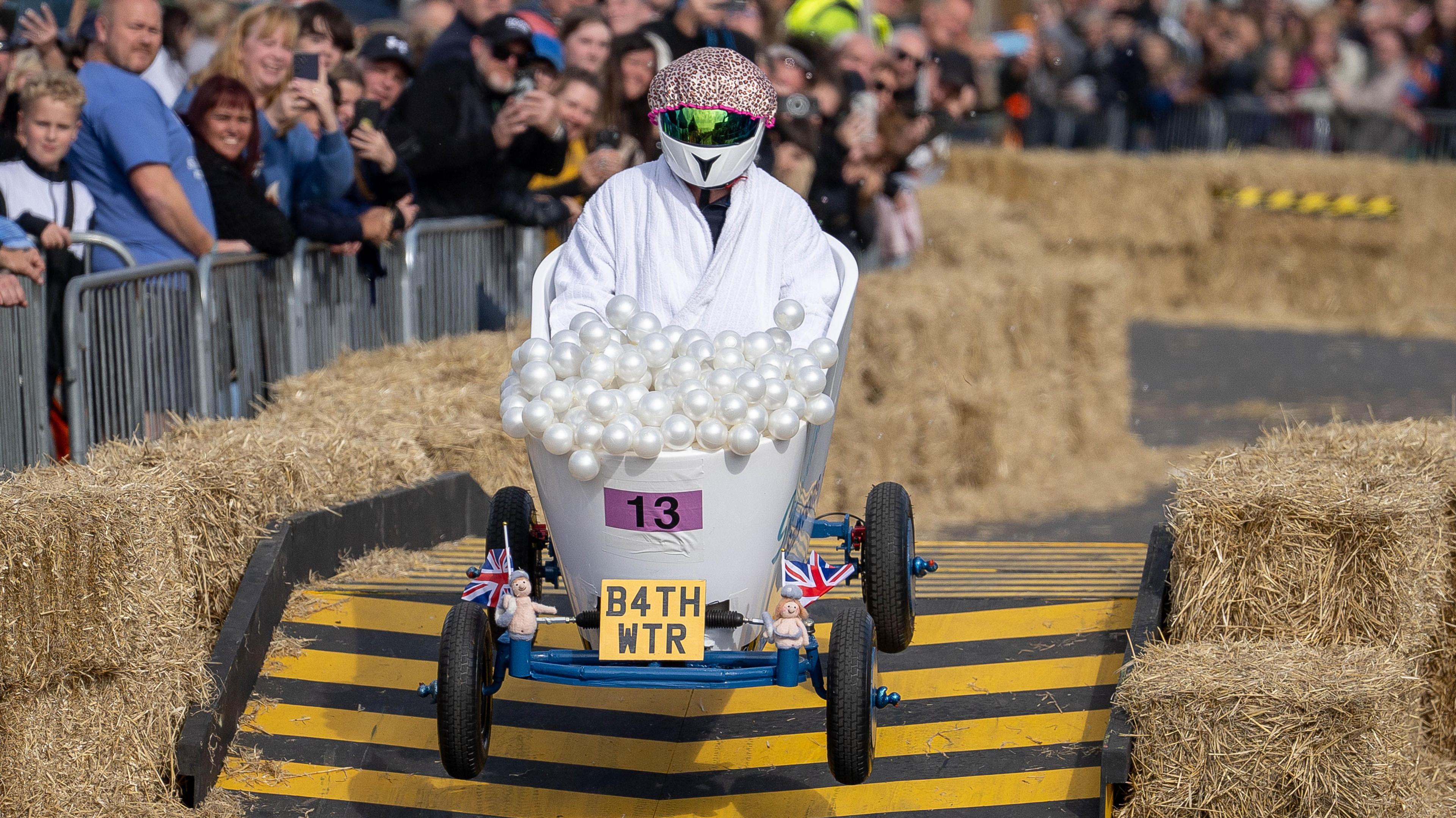A kart with a yellow number plate which uses letters and numbers to spell out BATH WATER. There are balls painted to look like bubbles on the top of the kart. It has been designed to look like a bubble bath. The driver is wearing a helmet with a leopard print shower cap on top.
