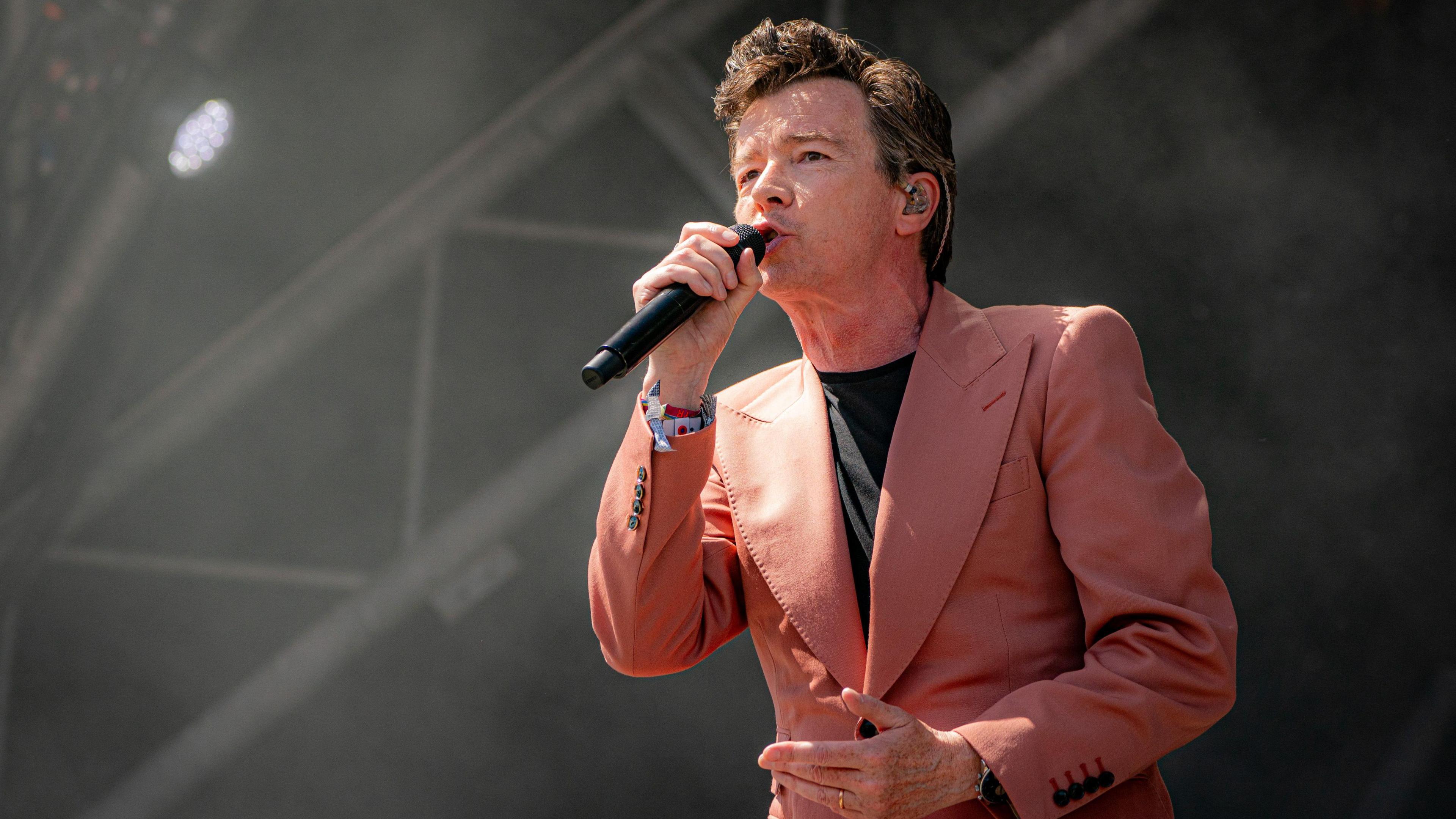 Rick Astley performing on the main stage at Glastonbury Festival in 2023, singing into microphone with pink jacket and black t-shirt 