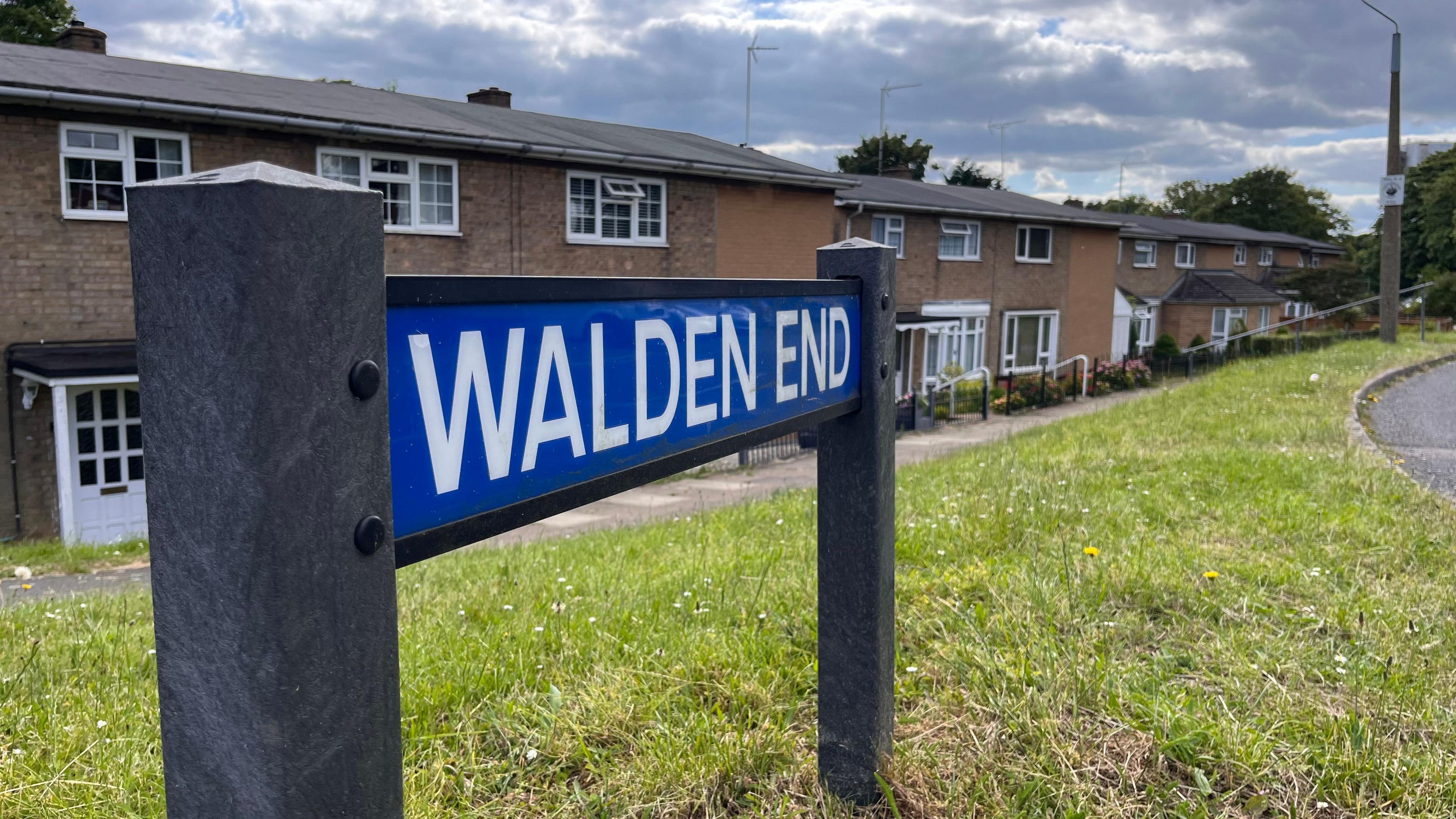 A blue road sign, attached to grey posts, saying "WALDEN END" in white capital letters. A row of light brown brick houses, with white windows, is in the background. 