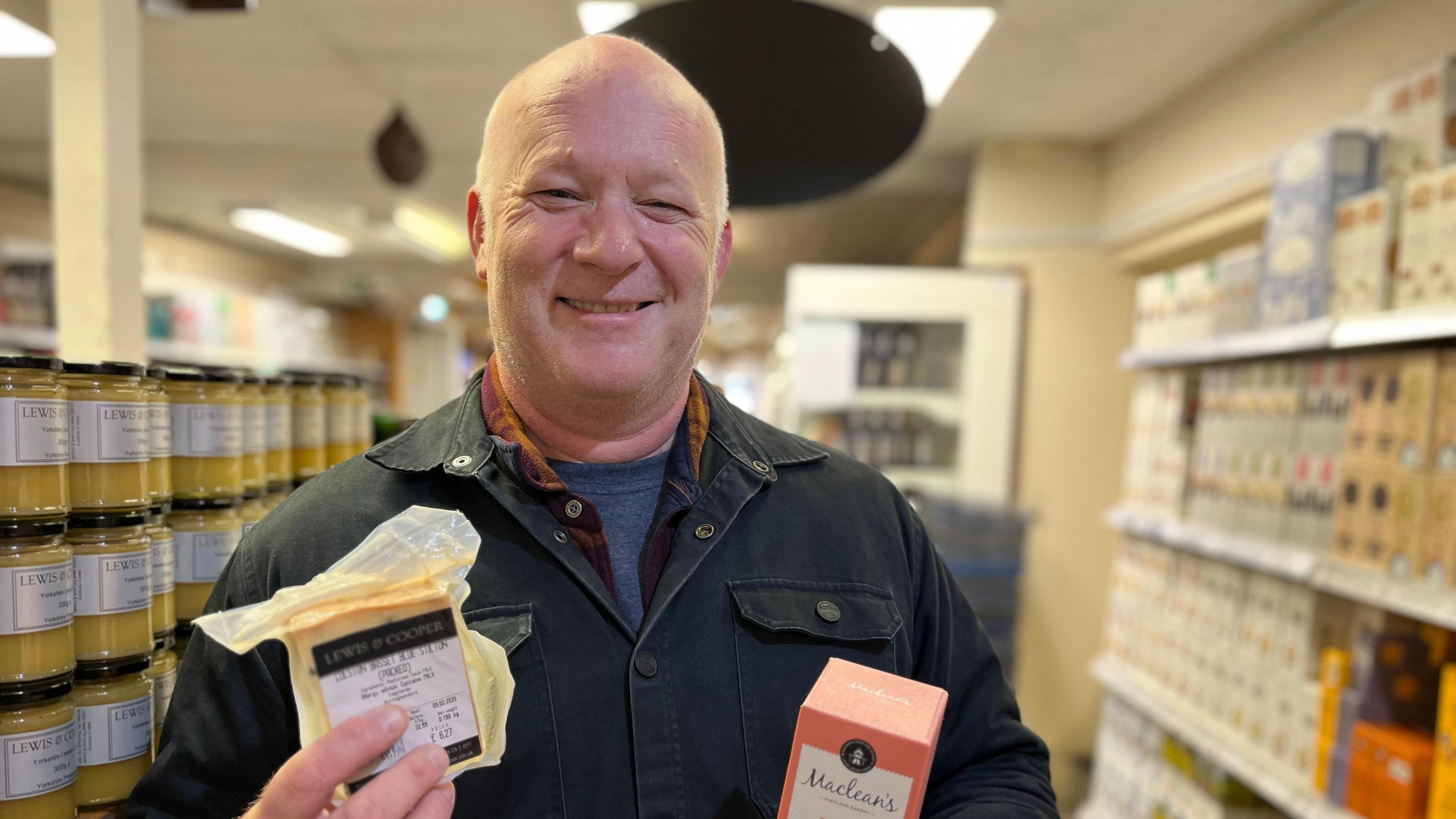 A bald man wearing a black jacket is standing in front of jars and packets of food.  He is holding up a packet of cheese and a packet of crackers.