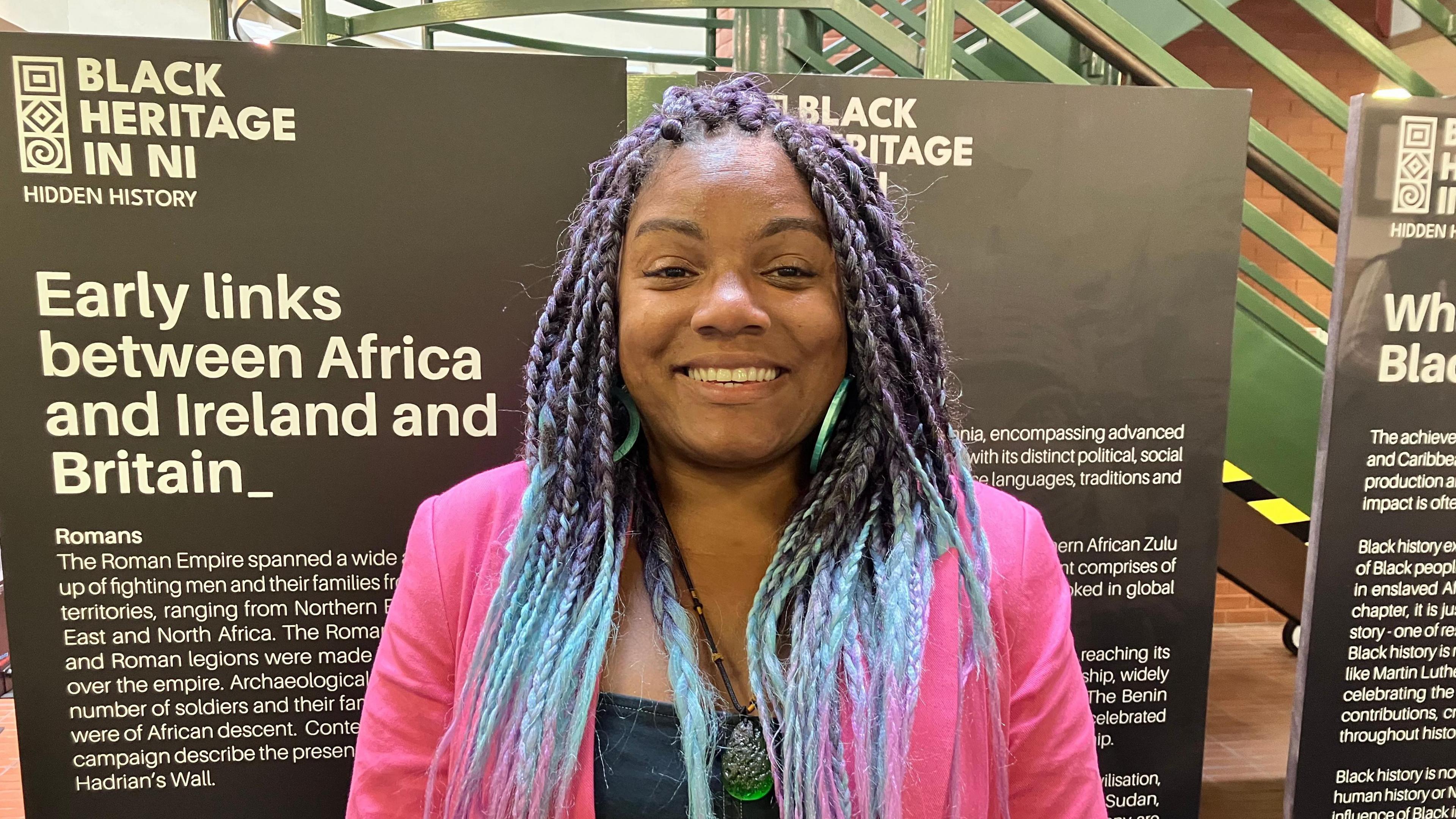 Woman has long and colourful blue and purple hair. 
She is wearing a bright pink jacket and green hoop earring and is smiling. 
She is standing in front of placards for the Black Heritage NI project which are positioned in front of a green staircase. 