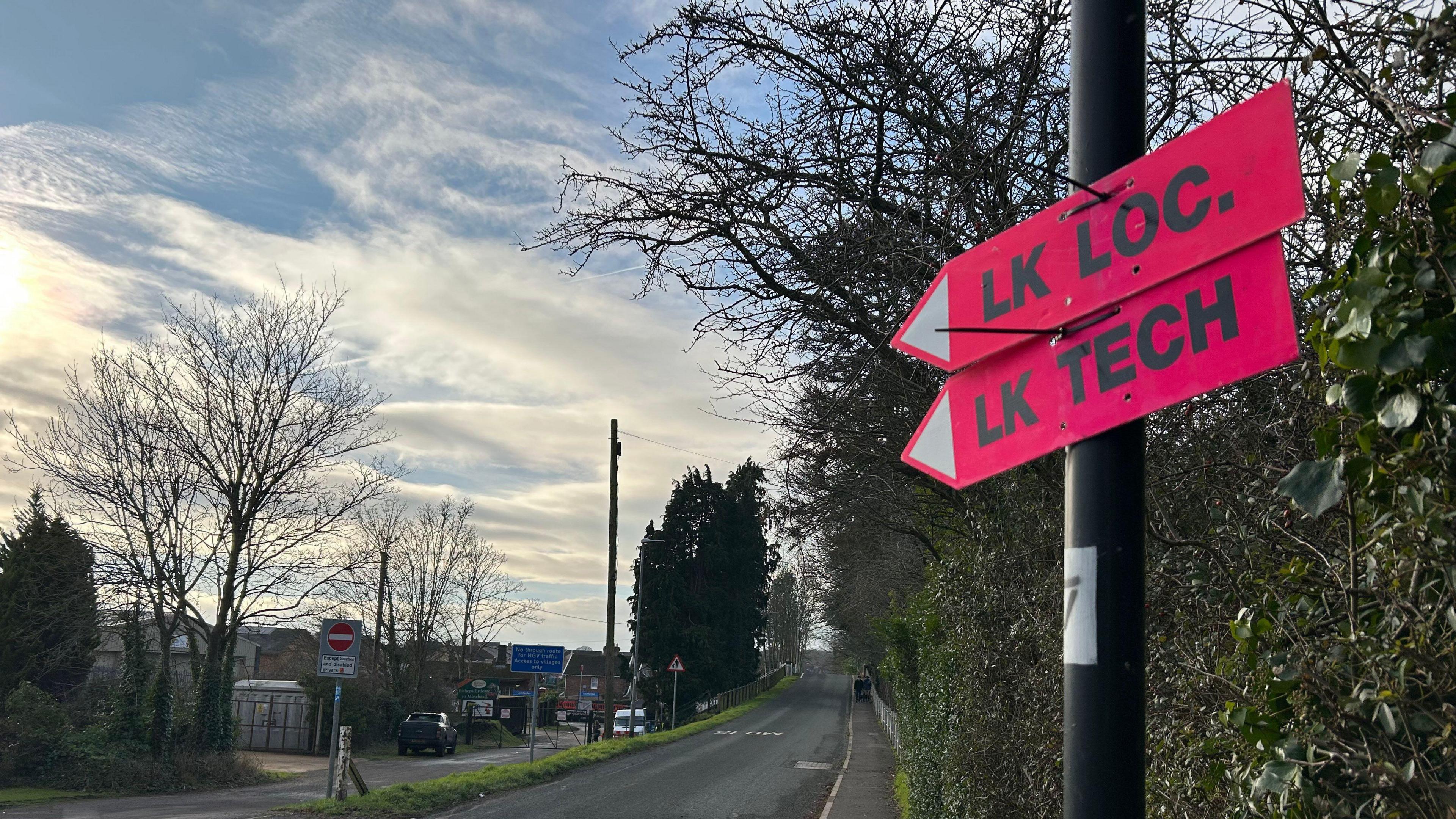 Two bright pink signs in the shape of arrows are stuck on a lamppost pointing left. One has 'LK Loc' written on it and the other has 'LK Tech' written on it. A road can be seen in the background. 