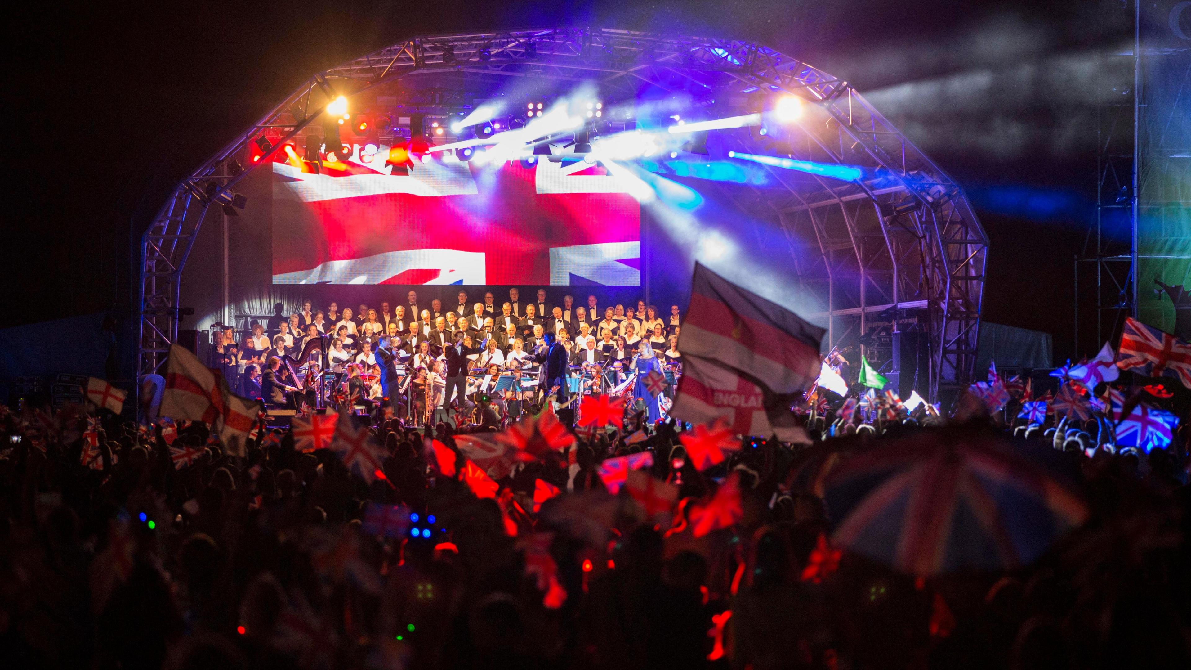 Bedford Proms in Bedford Park, with an orchestra and singer on the stage, the Union Flag behind them, and crowd members, waving their flags. Lights are above the stage.