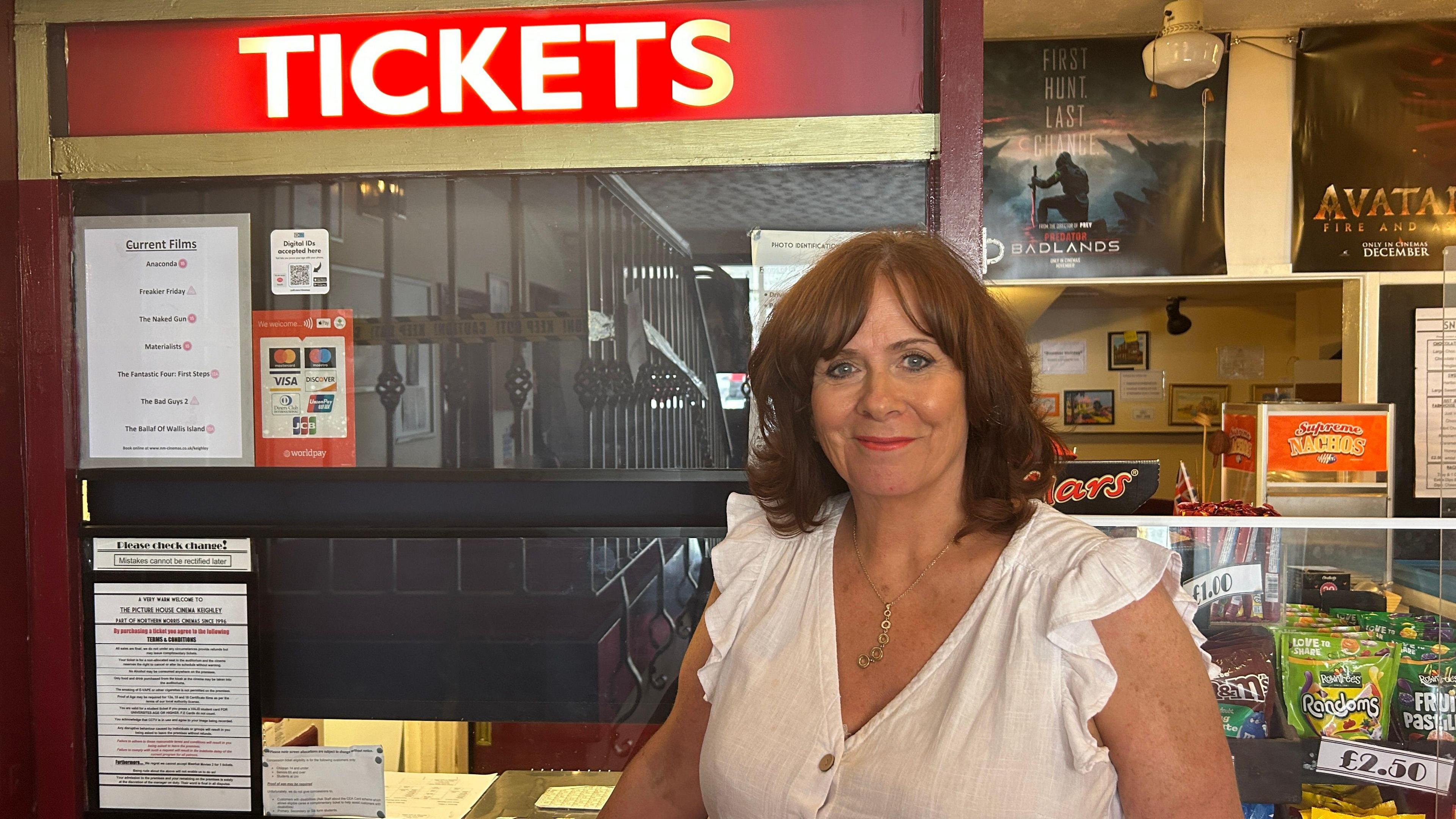 A middle aged women standing next to a cinema ticket and snacks kiosk. Film posters, the ticket window and shelves of sweets can be seen behind her.