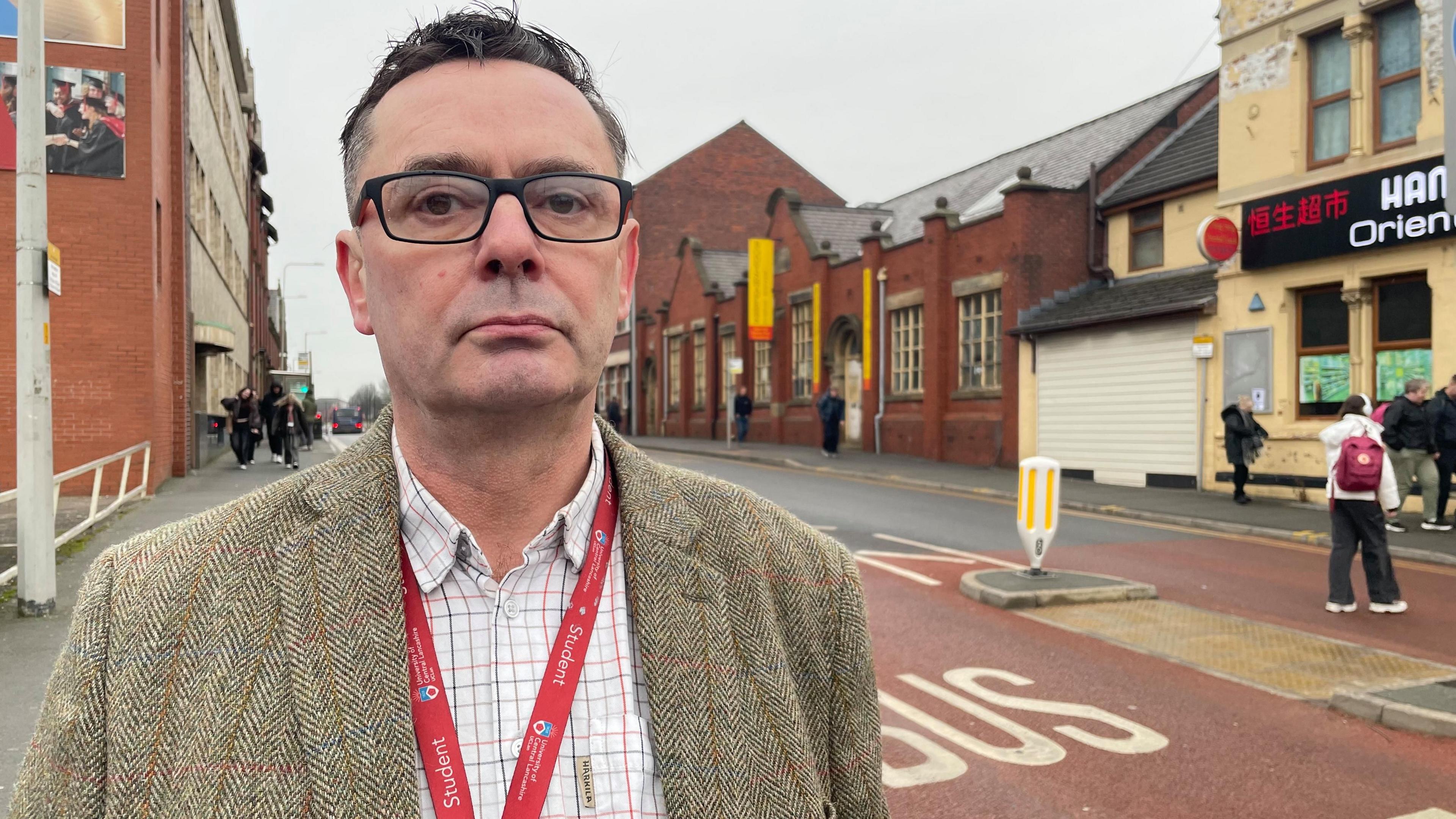 Chris Fisher standing in front of the 'bus gate' sign on the road.  The road says 'bus gate' in white on a red background on the left hand side.  People are walking past a Chinese restaurant on the other side of the road.  In the background there are red brick buildings and people walking towards Chris.