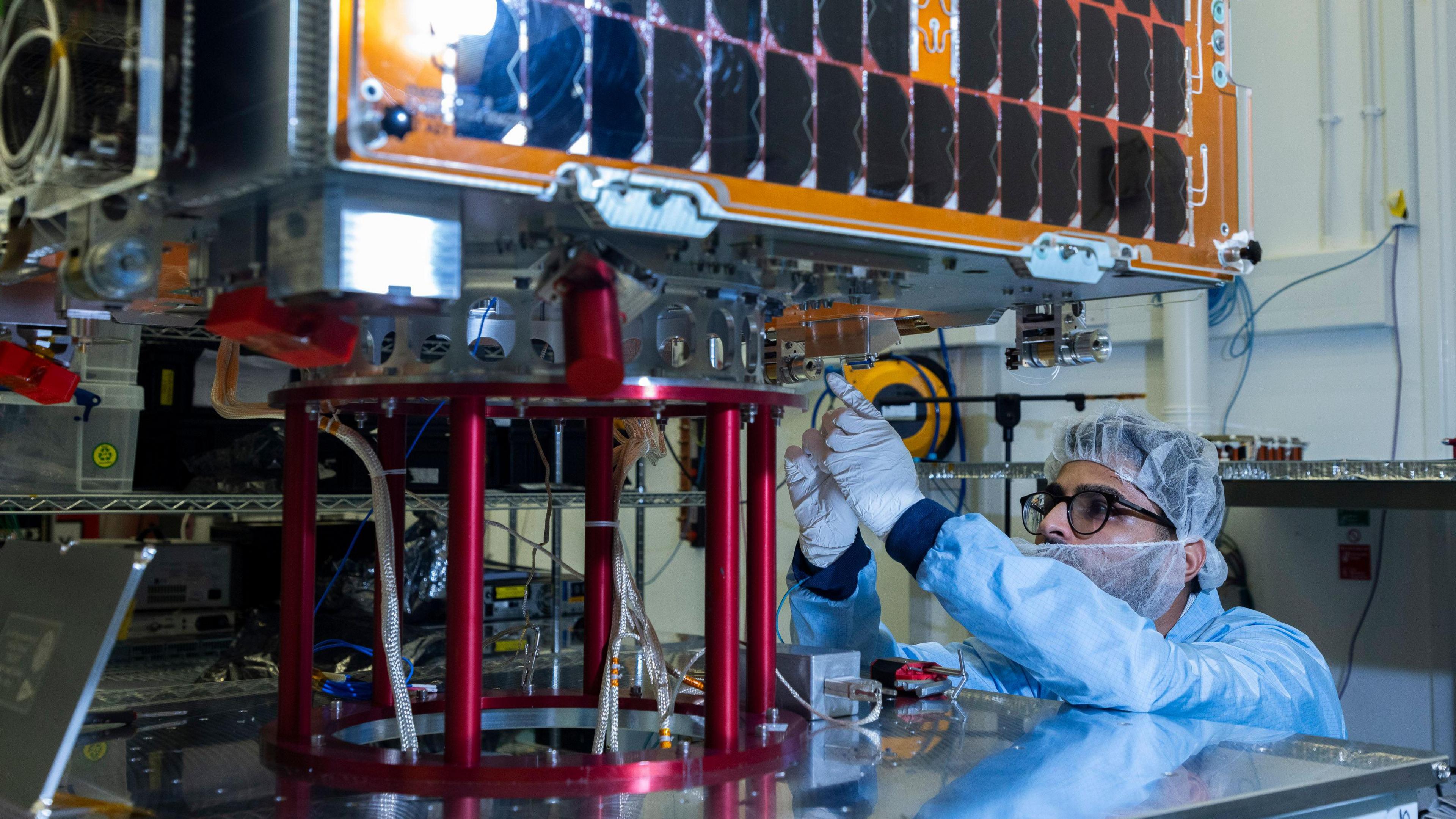 A man wearing protective gear fixes the Tyche satellite in a laboratory.

