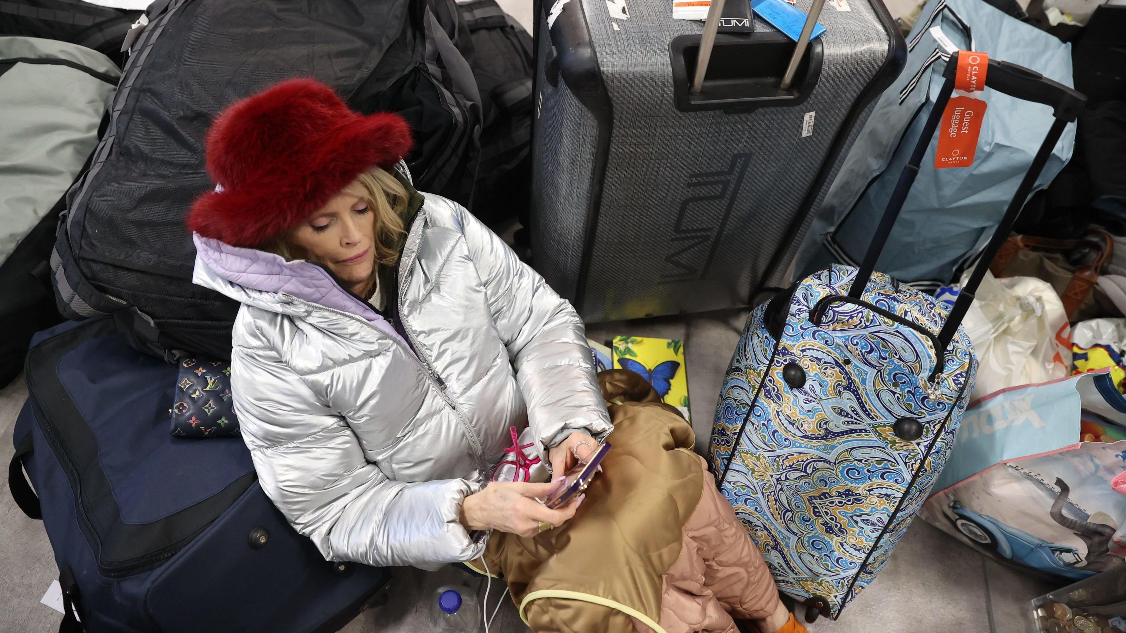 A woman, wearing a silver coat and a red hat, sits among a pile of rucksacks and suitcases as she looks at her phone