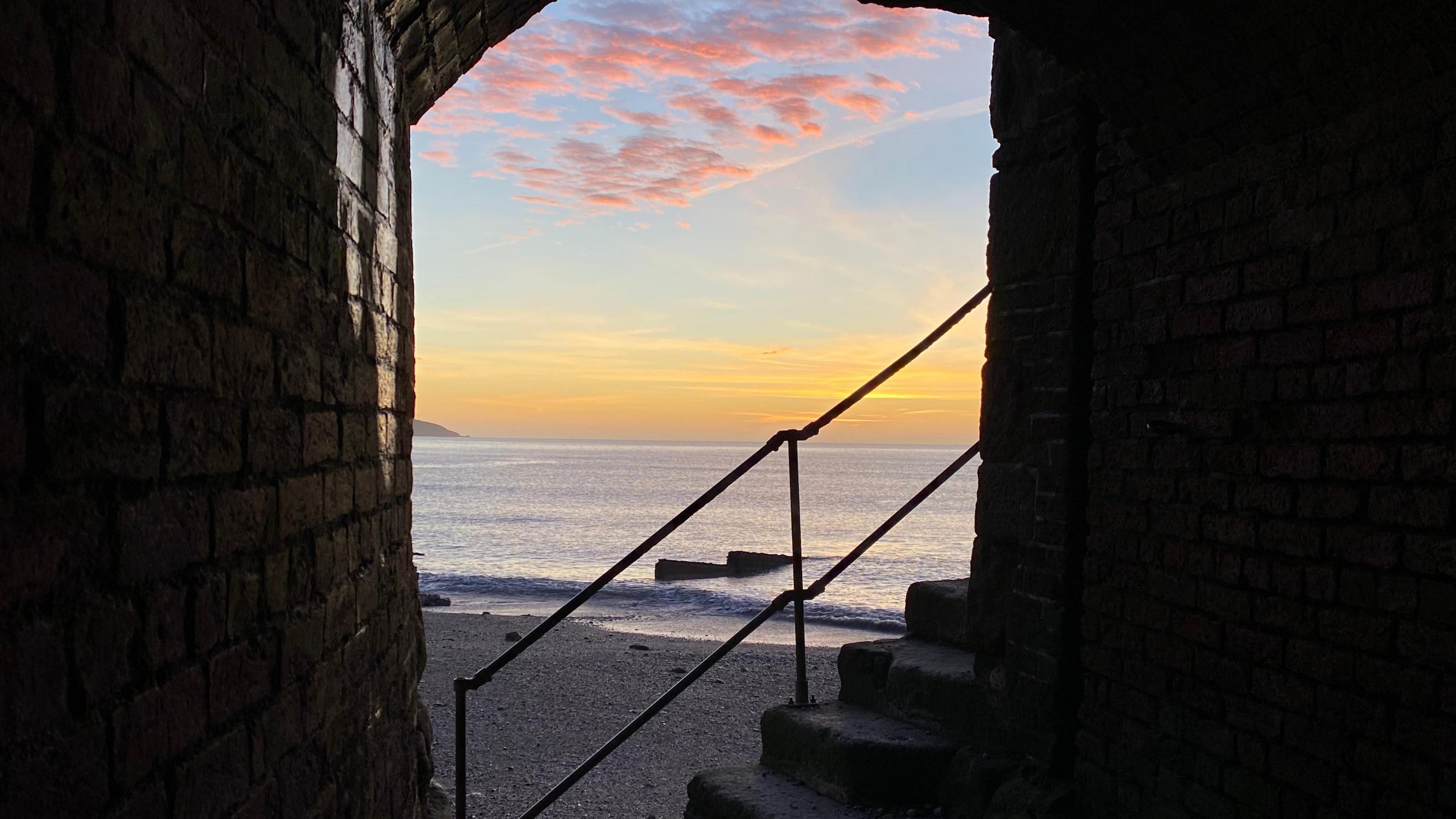 Clouds with a pink hue are in the sky above Charlestown. Small waves are heading into the beach. The walls of a structure and stone steps with metal railings next to them. The sky has blue and yellow colours.