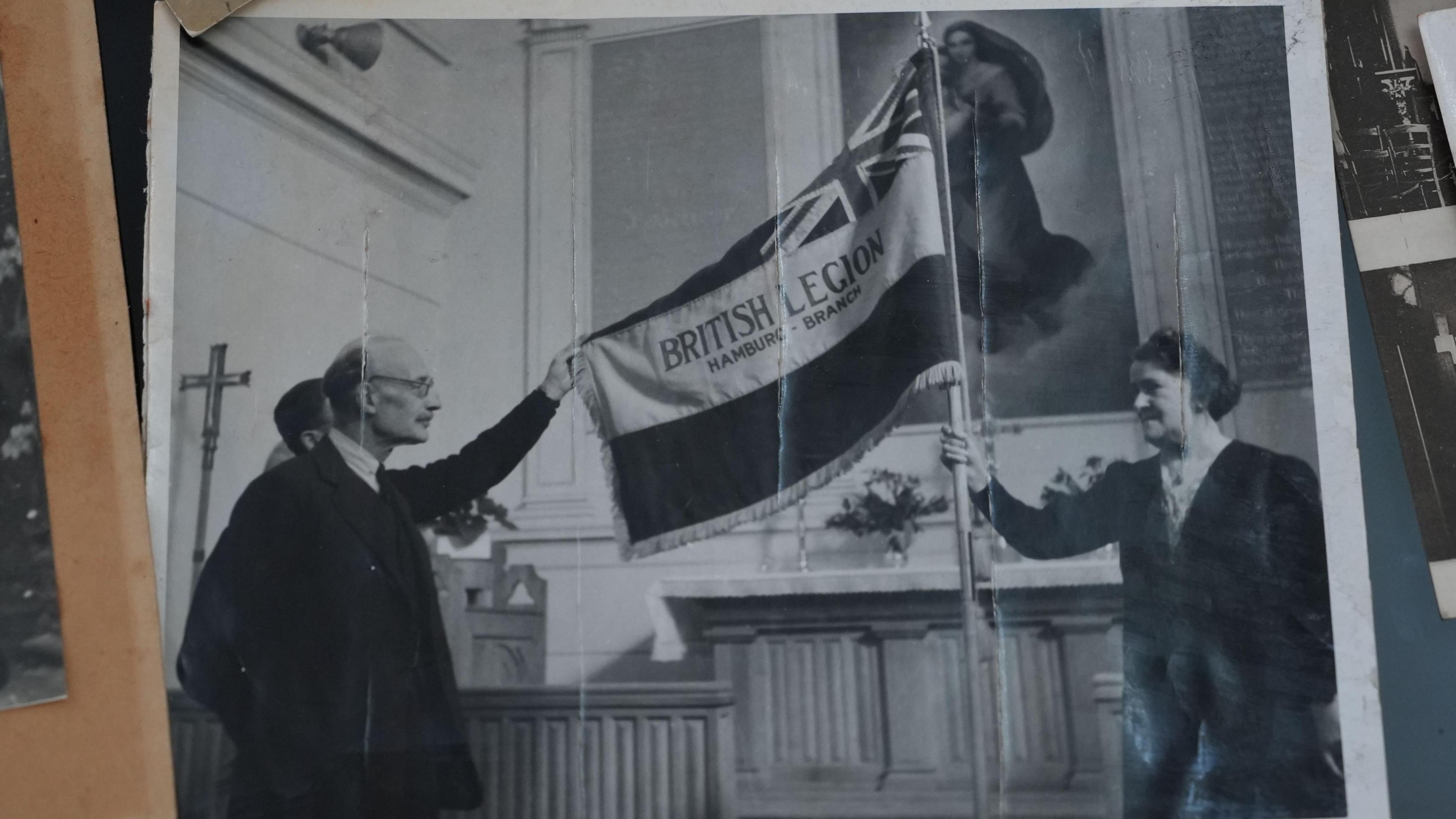 In another black and white photograph, Mabel Wulff is pictured on the right holding the British Legion flag aloft in the English Church in Hamburg. Pictured with two men in suits, the trio are holding the flag up in front of the altar. 