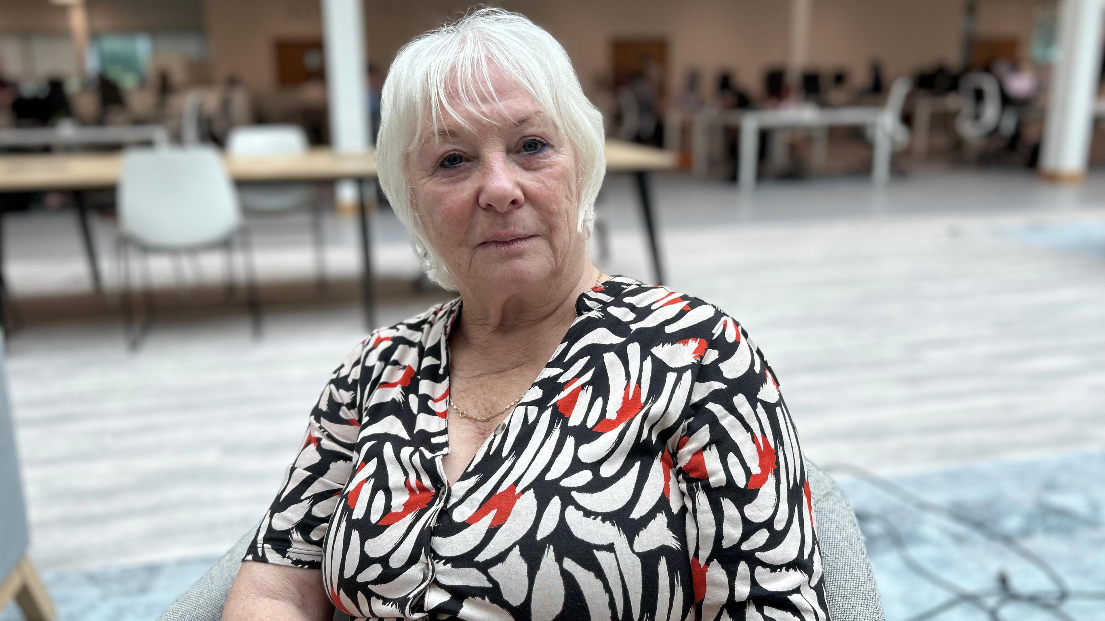 Woman with white hair and black, white and red top. She is sitting in a hall. In the background are tables and chair.