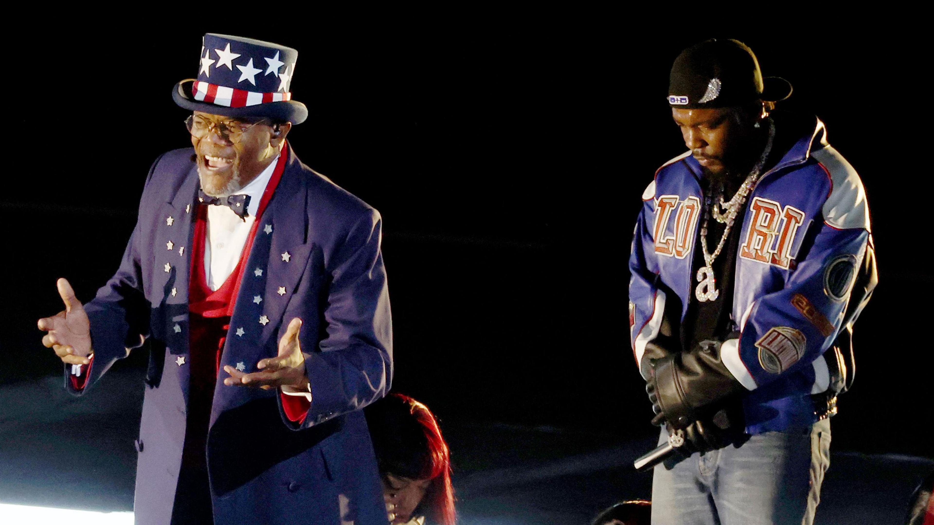 Samuel L Jackson as Uncle Sam during Kendrick Lamar's halftime show. Jackson wears blue tails with stars on the lapels over a red waistcoat, white shirt and black bow tie. His outfit is completed by a blue top hat decorated with the stars and stripes of the US flag. He holds his hands out, mid speech, while Kendrick stands behind him, looking down with his bands folded in front of him. 